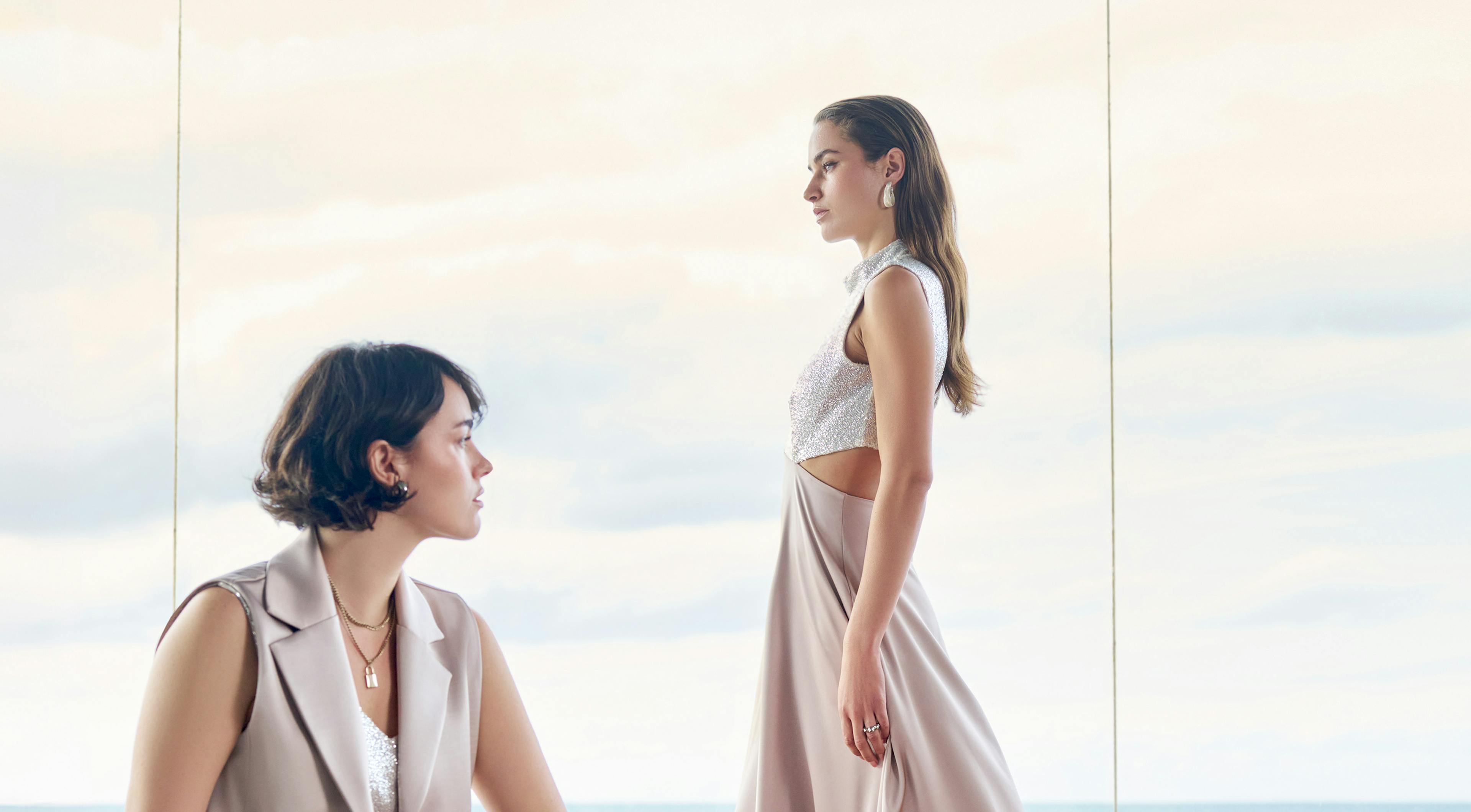 Two women in elegant dresses are posed by a large window with a cloudy sky behind them. One sits gazing forward; the other stands in profile.