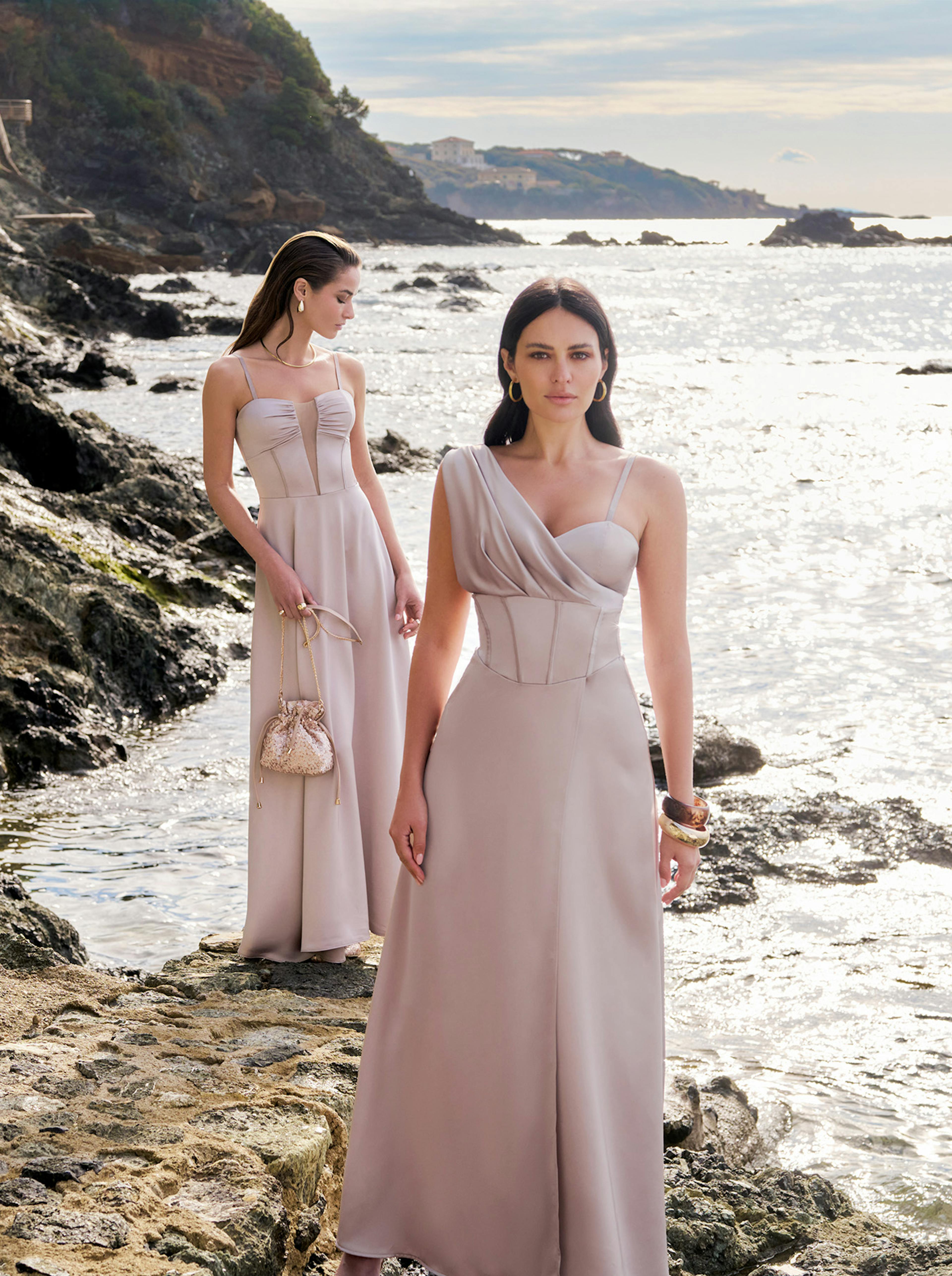 Two women in elegant, flowing mauve dresses stand on rocky shore by the sea. One gazes ahead confidently; the other looks aside, holding a purse. The scene is serene and sophisticated.