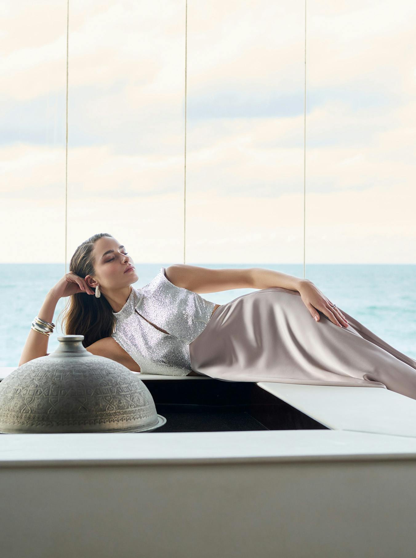 A woman in a shimmering silver top and flowing skirt lounges elegantly by a window overlooking the ocean. The setting is serene and luxurious.