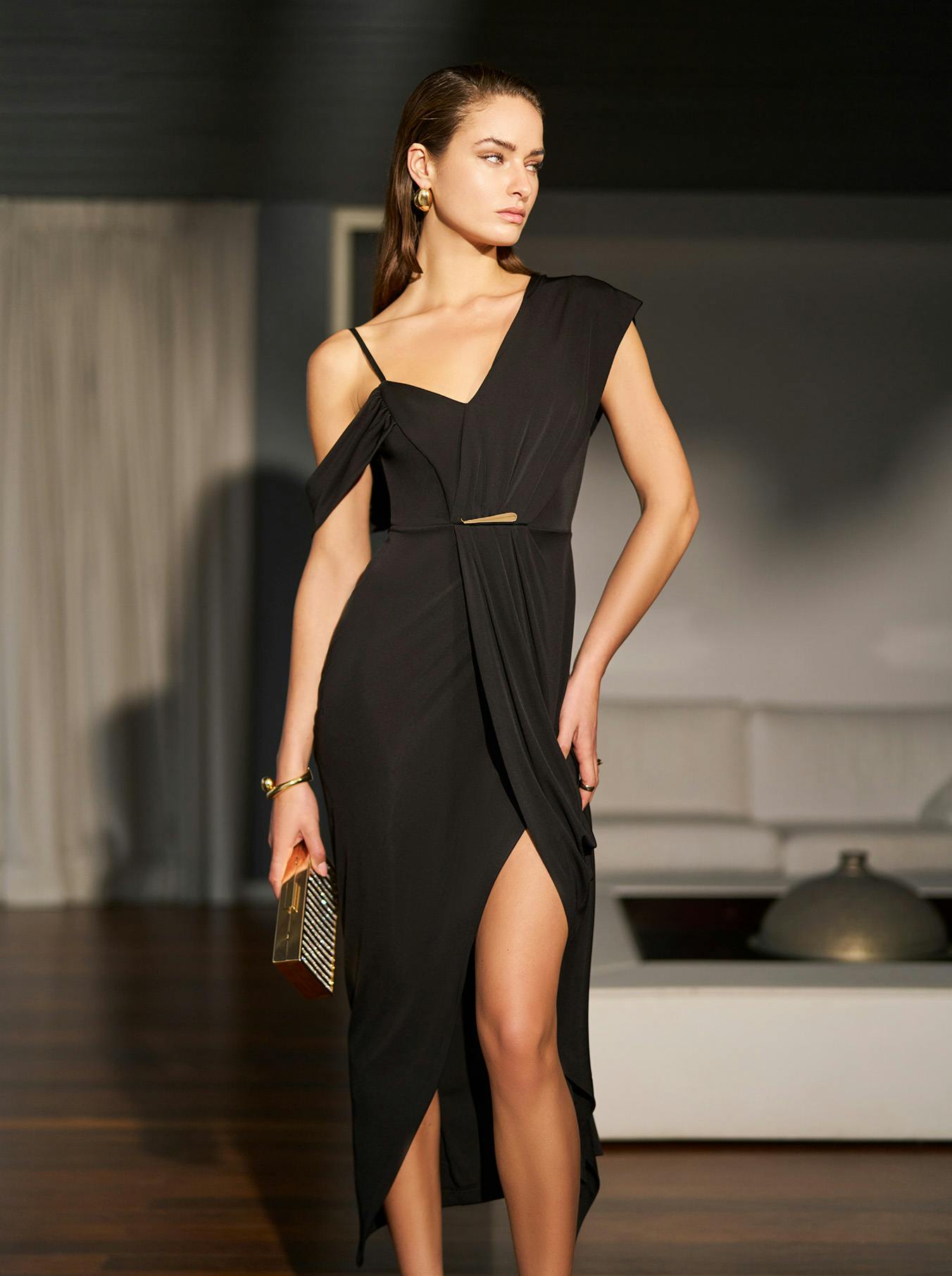 A woman in a black, one-shoulder dress with a thigh-high slit stands confidently in a modern room. She holds a gold clutch, exuding elegance.