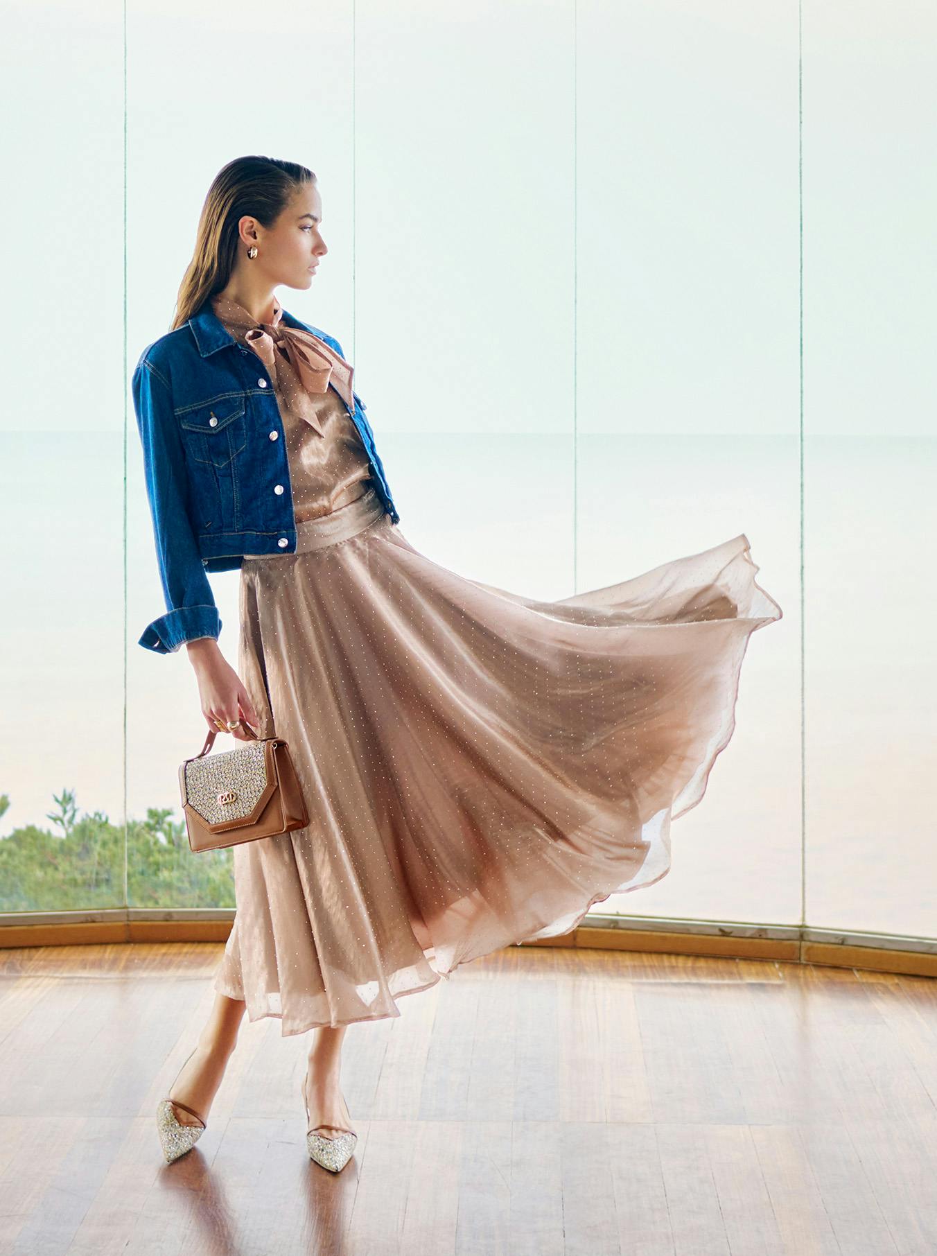 Woman in a flowy taupe dress and denim jacket stands gracefully, holding a small handbag. Backdrop of large windows with a serene ocean view.