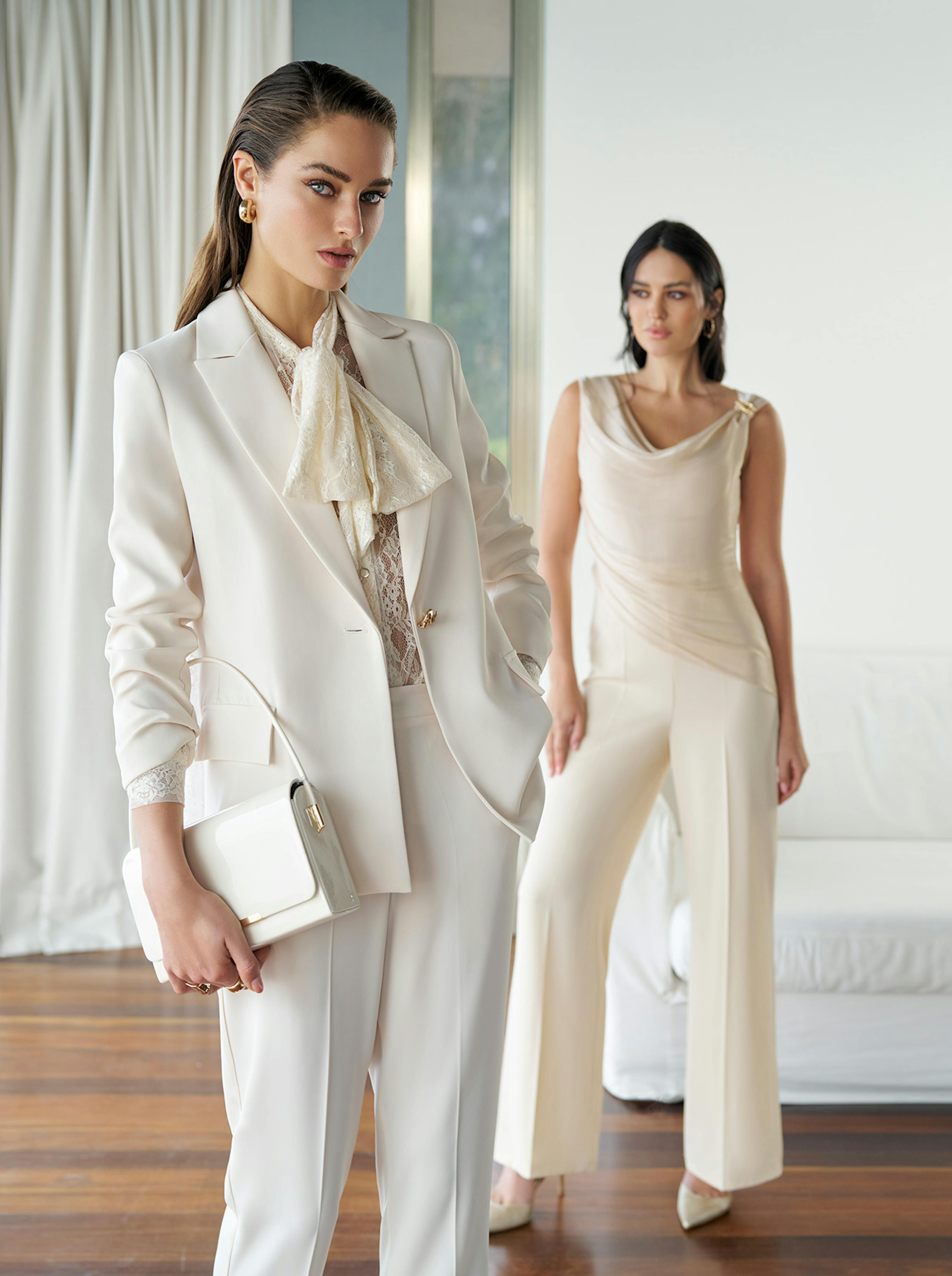 Two women in an elegant setting. One in a beige suit and lace blouse with a scarf and handbag, the other in a creamy draped top and pants. Sophisticated and poised.