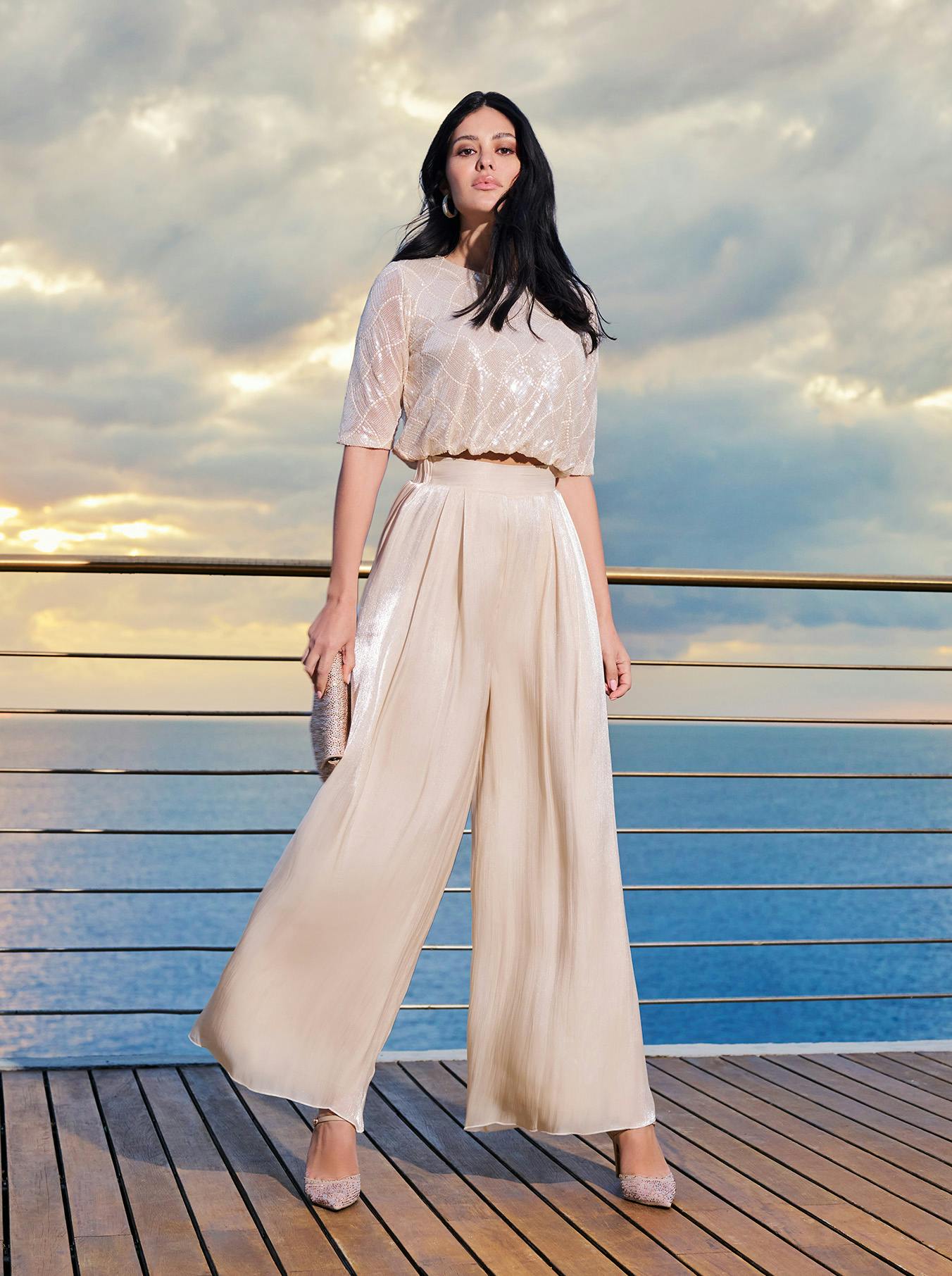 Woman in a shimmering, loose-fitting beige outfit stands on a wooden deck. A deep blue sea and cloudy sunset create a serene backdrop.
