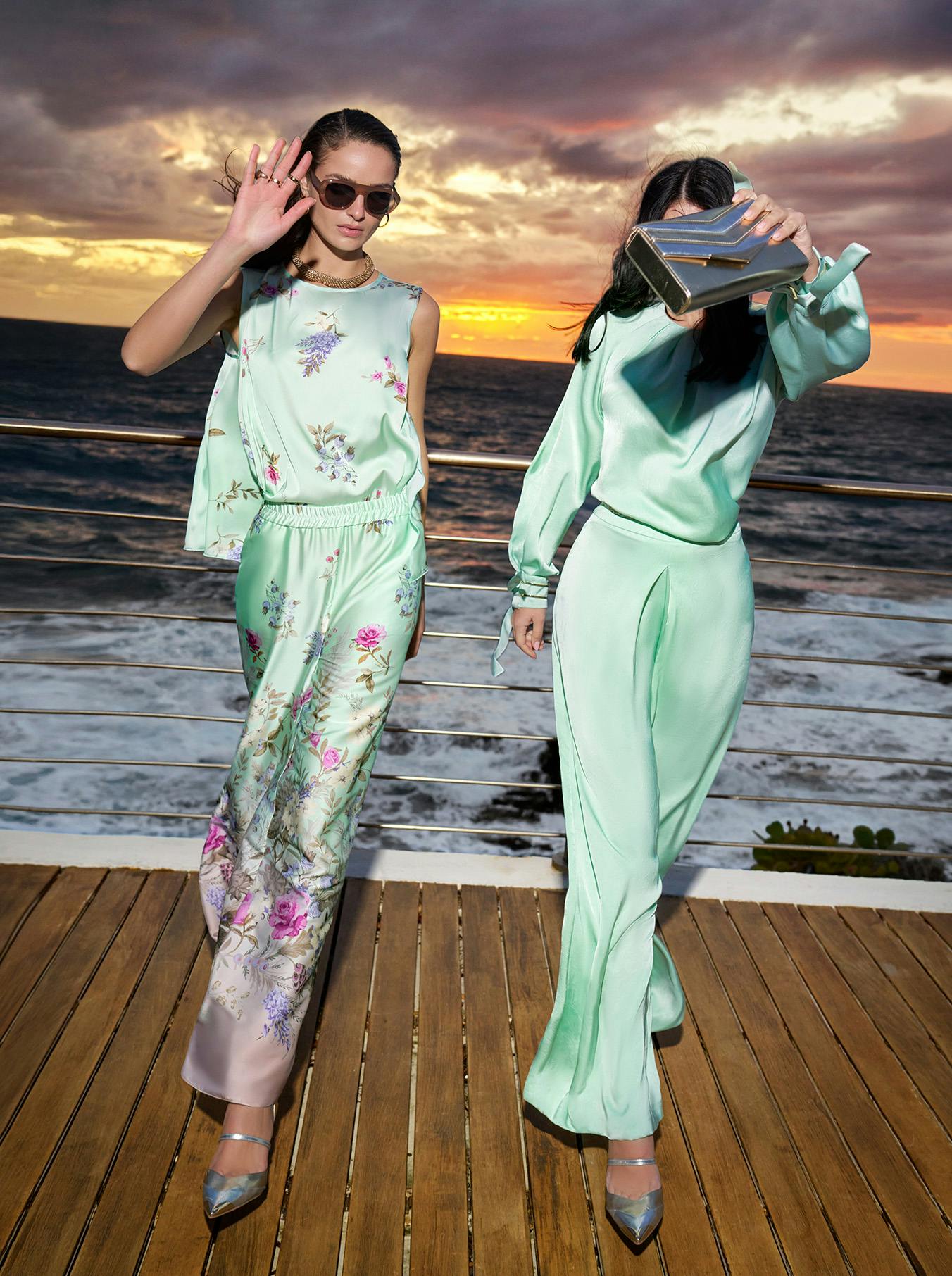 Two women in pastel floral and green outfits pose on a wooden deck with a dramatic ocean sunset background. One waves, the other covers her face with a clutch.