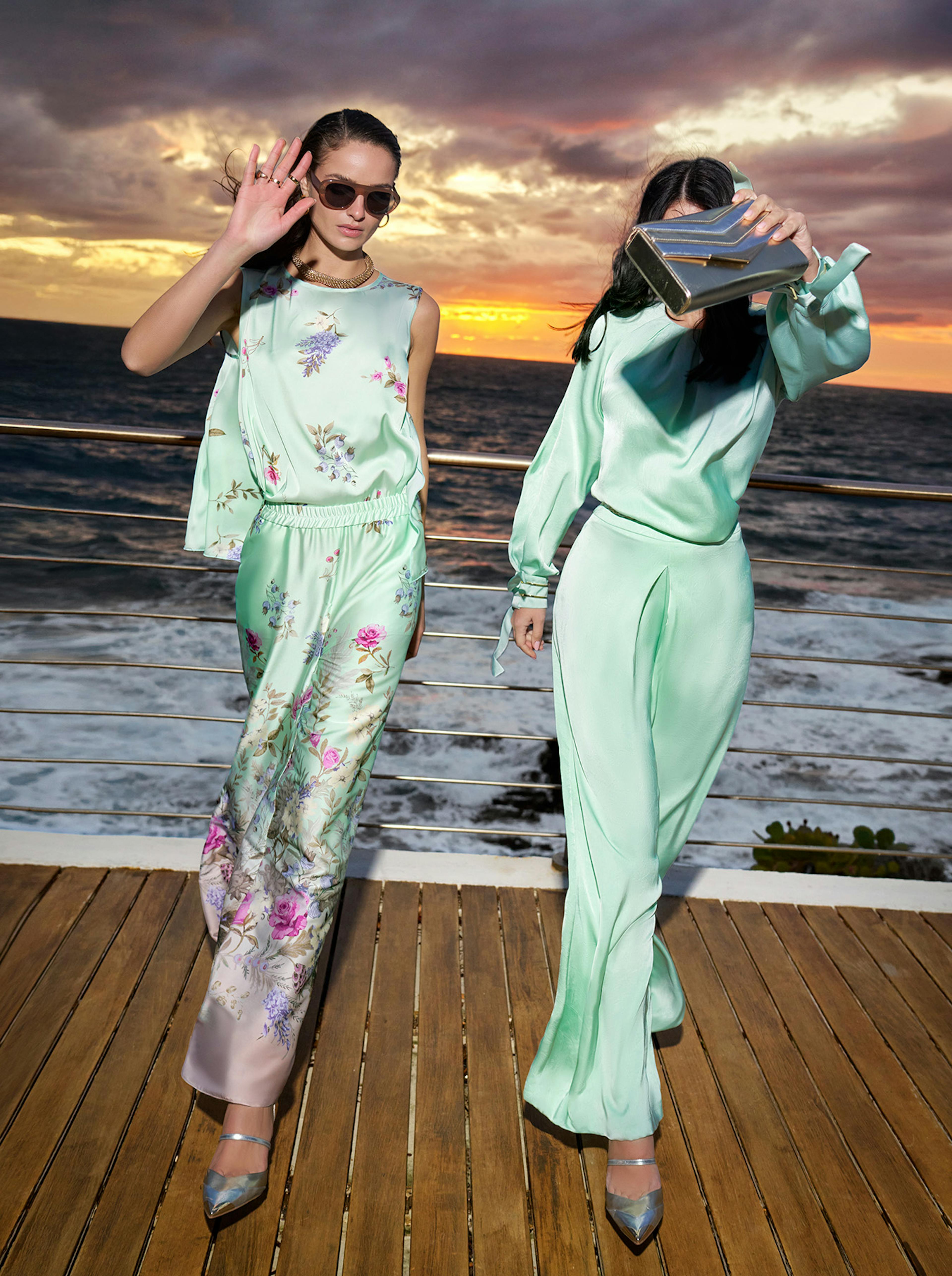 Two women in pastel floral and green outfits pose on a wooden deck with a dramatic ocean sunset background. One waves, the other covers her face with a clutch.
