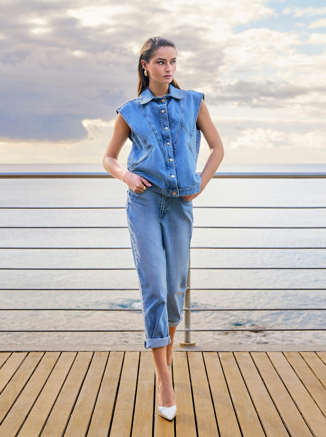 A woman in denim clothing stands on a wooden deck by the sea, hands in pockets. The cloudy sky and serene ocean create a calm mood.
