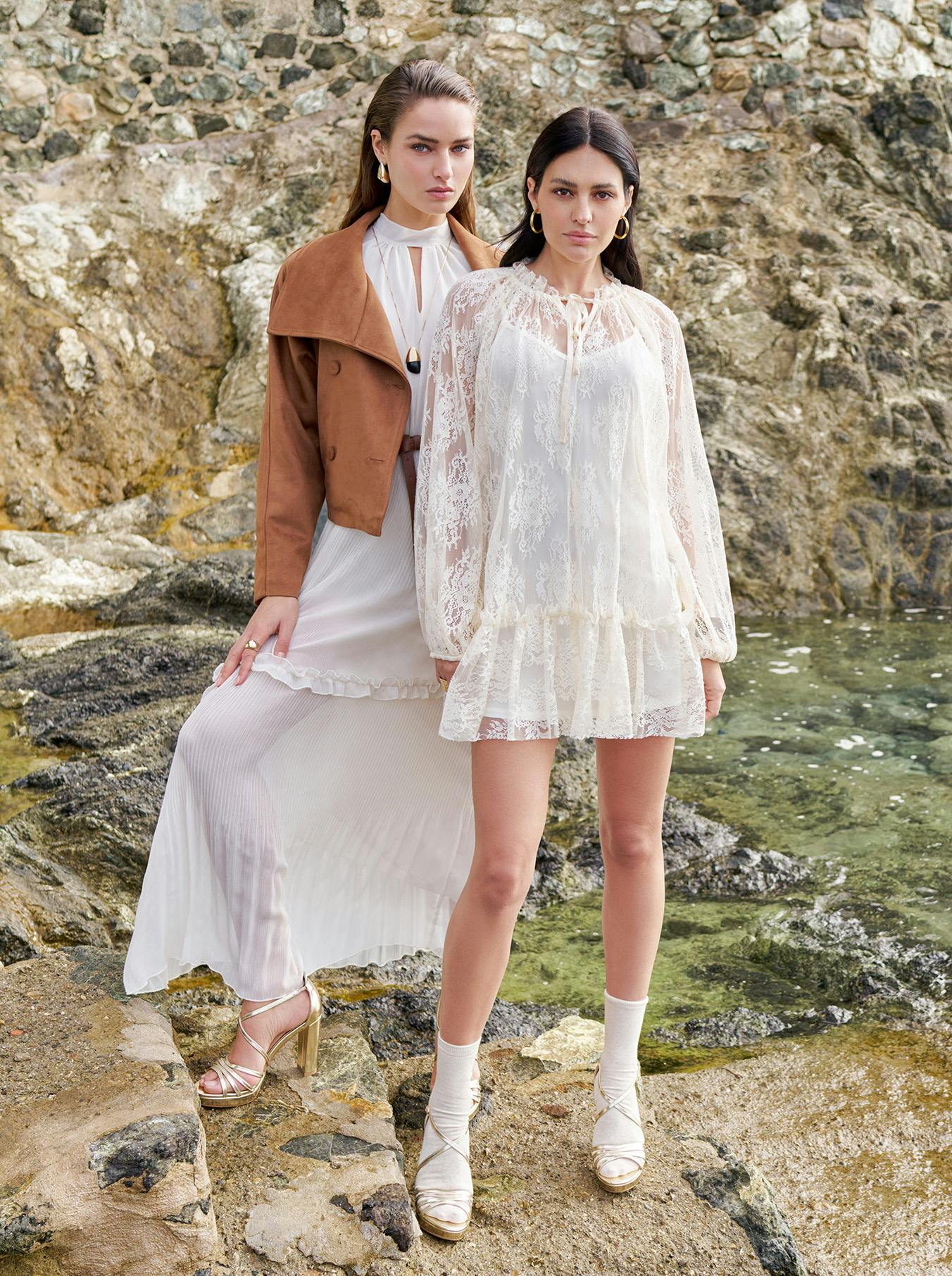 Two women in elegant outfits stand on rocky terrain by the sea. One wears a white lace dress, the other in a white pleated dress with a brown jacket. The scene is stylish and serene.