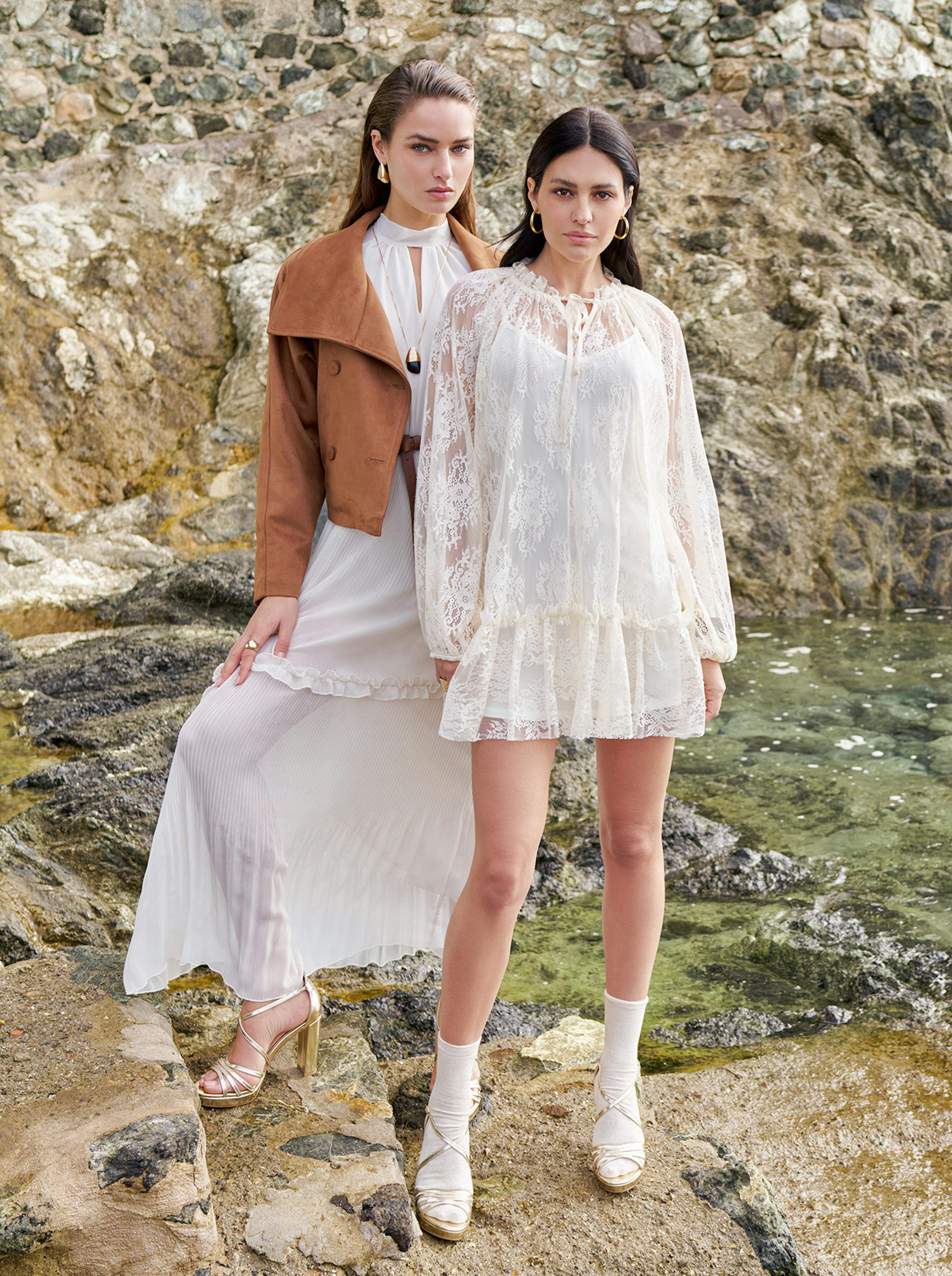 Two women in elegant outfits stand on rocky terrain by the sea. One wears a white lace dress, the other in a white pleated dress with a brown jacket. The scene is stylish and serene.
