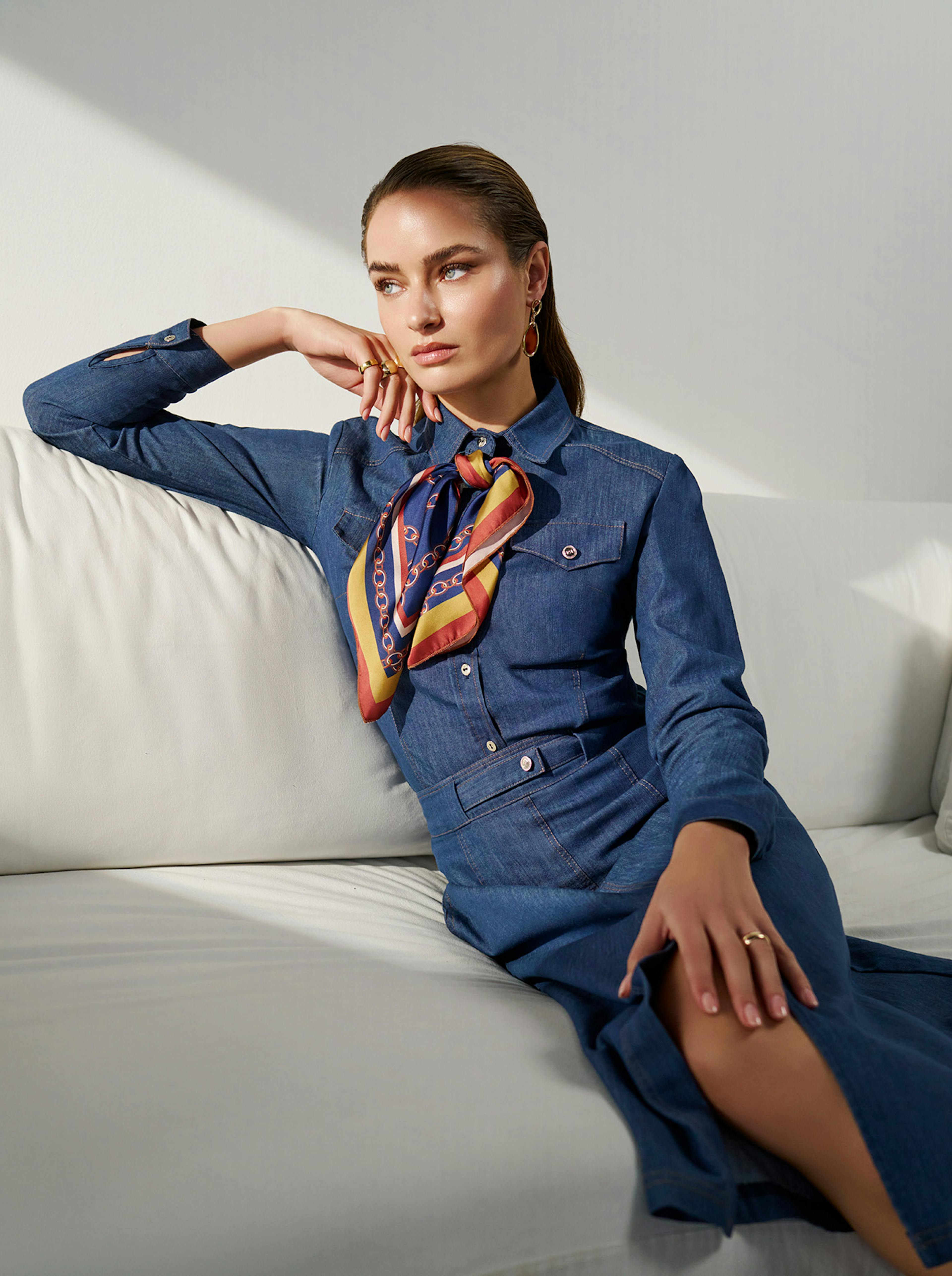 A woman in a denim dress and a colorful scarf sits on a white couch. Her expression is thoughtful, with soft lighting and a minimalist background.