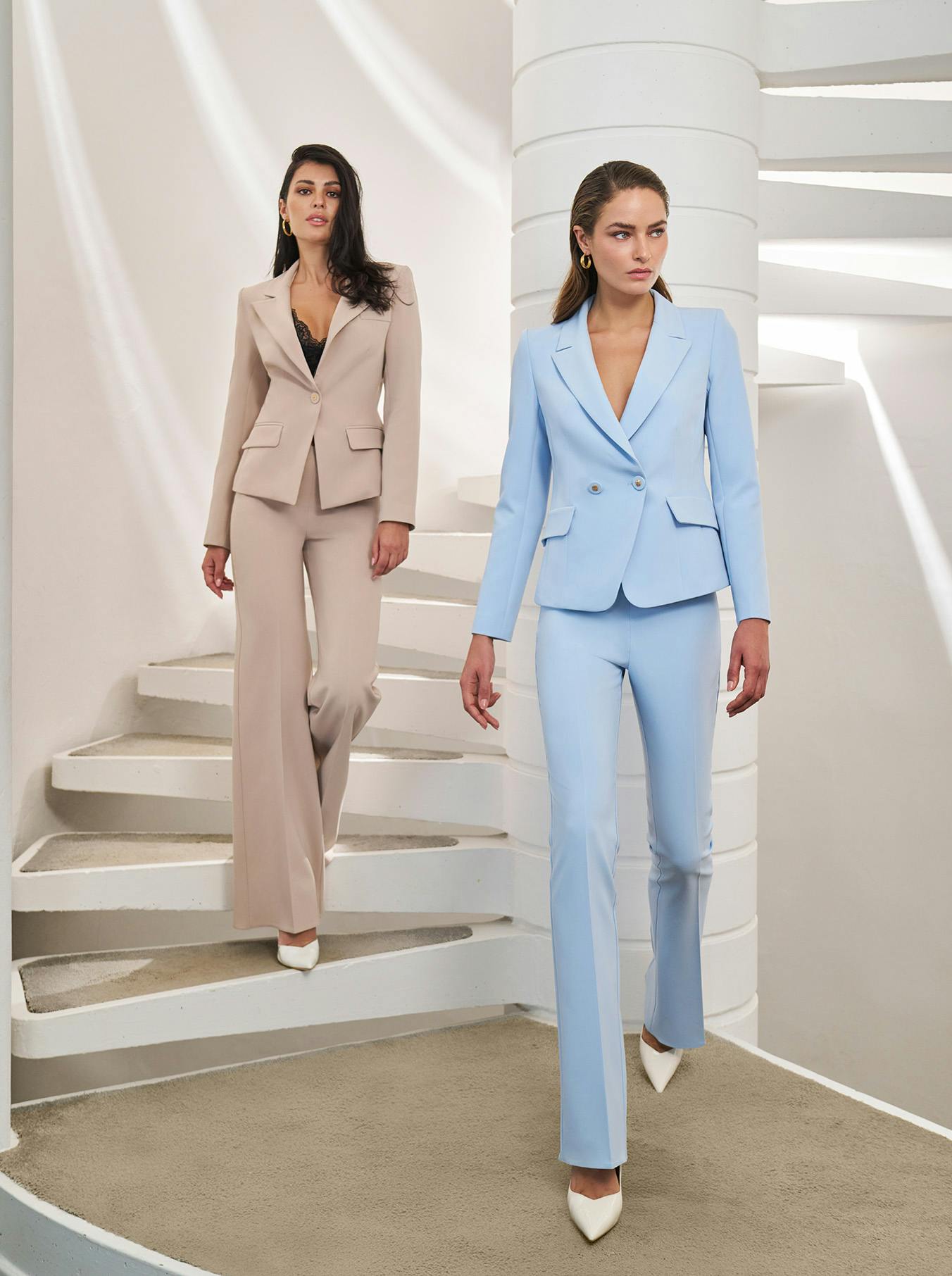 Two women confidently walk down modern white spiral stairs. One wears a beige suit, the other a light blue suit, both with white heels. Elegant and chic setting.