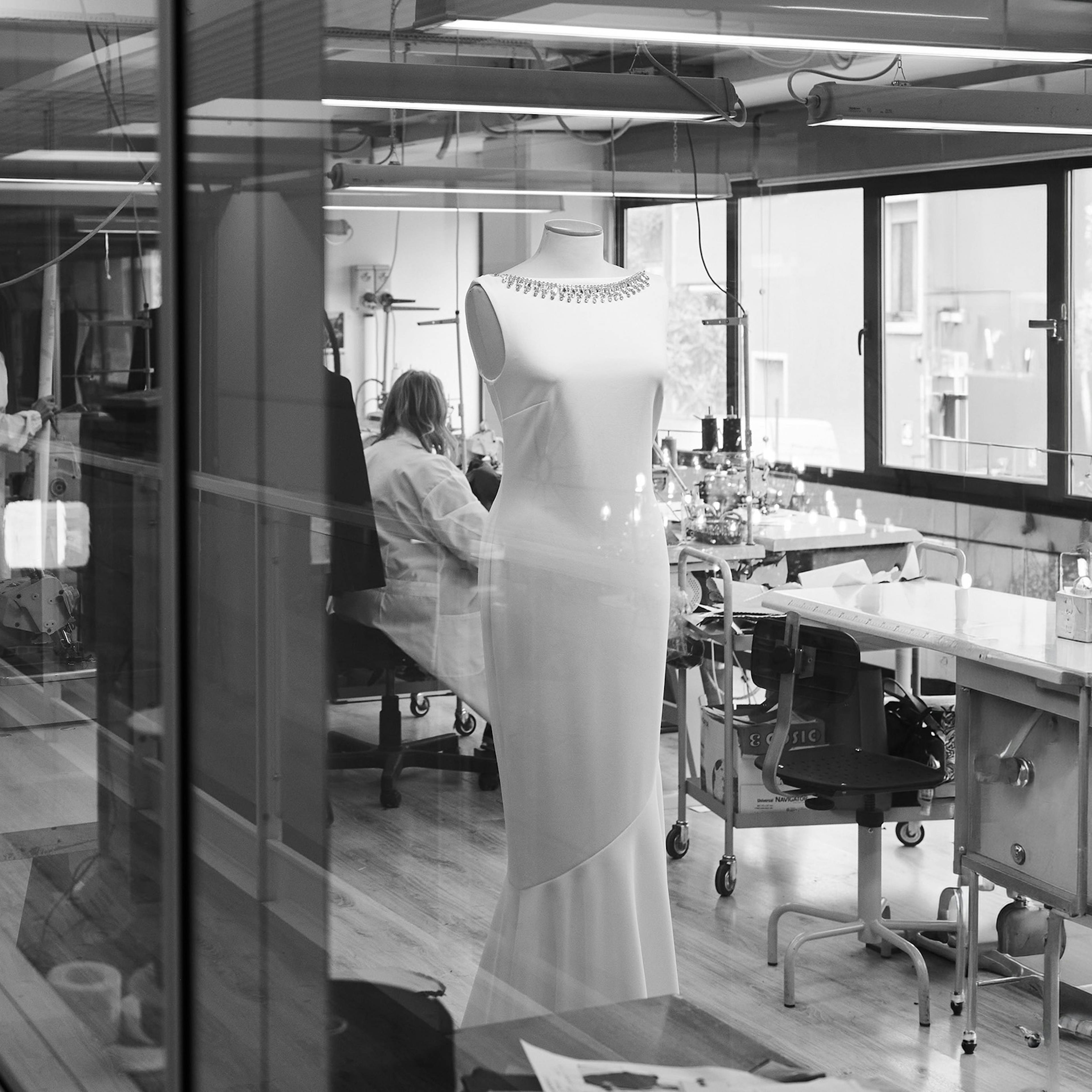 Black and white photo of a modern design studio. In the foreground, a mannequin displays an elegant dress. Designers work at cluttered desks in the background.