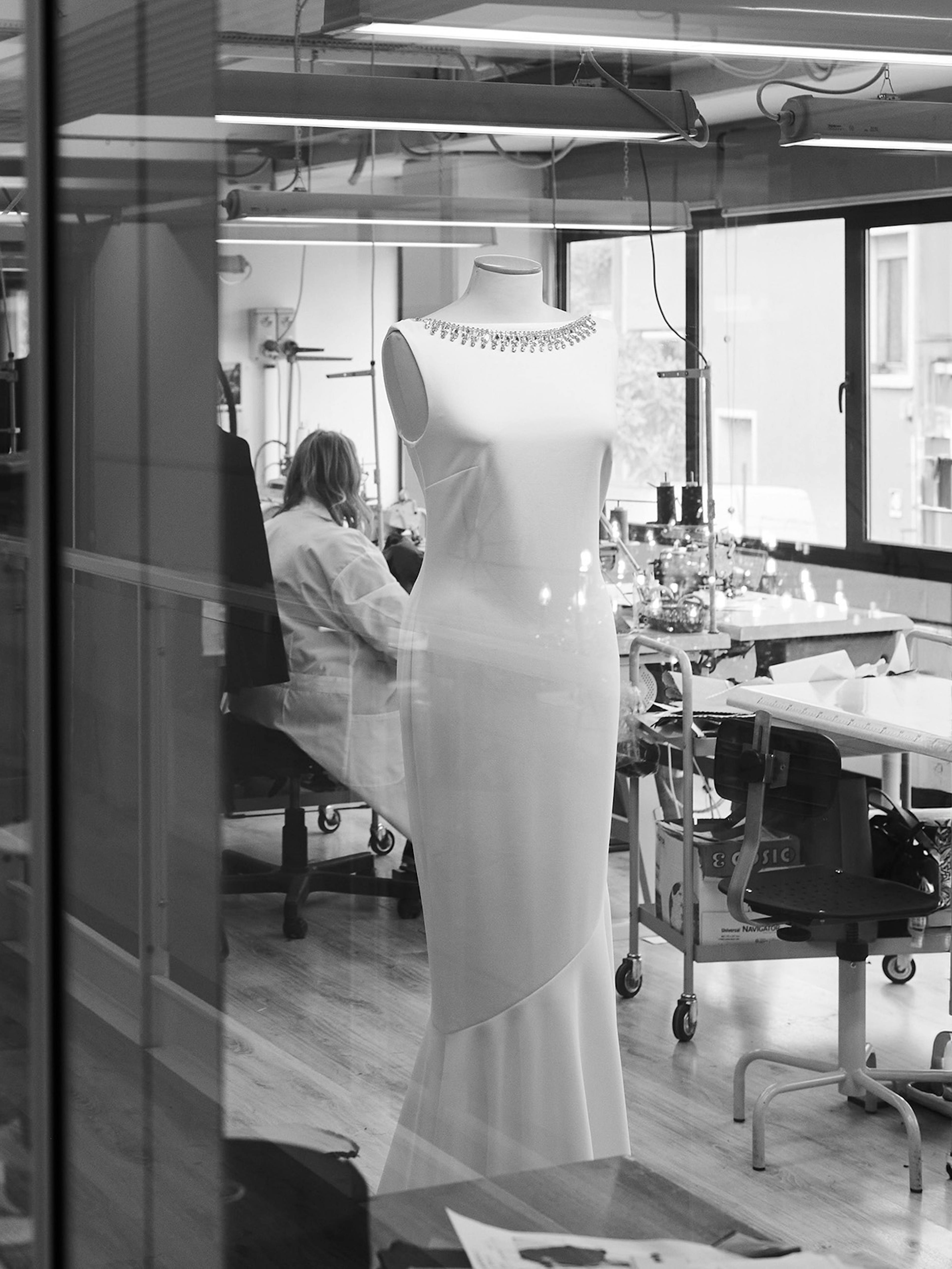 Black and white photo of a modern design studio. In the foreground, a mannequin displays an elegant dress. Designers work at cluttered desks in the background.