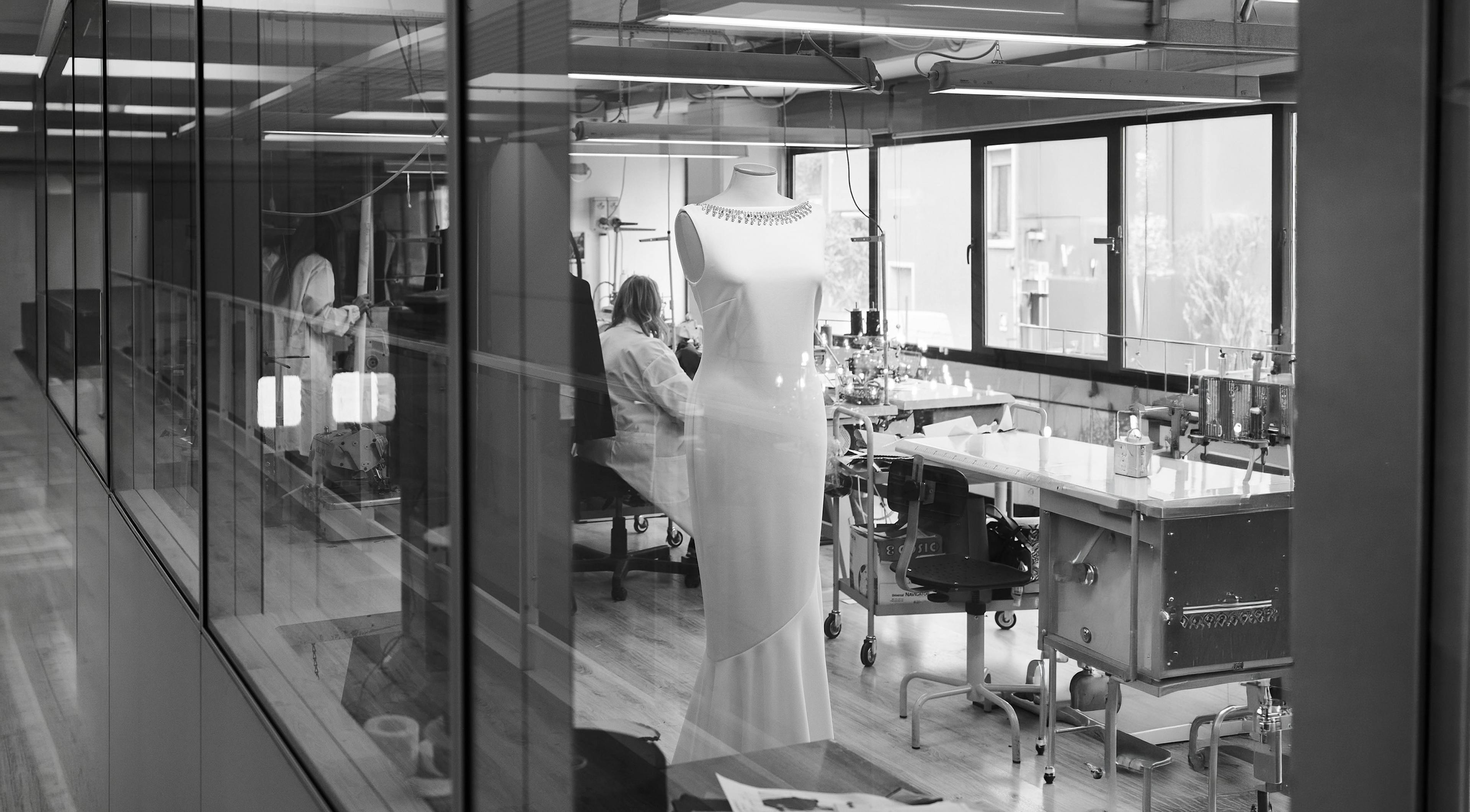 Black and white photo of a modern design studio. In the foreground, a mannequin displays an elegant dress. Designers work at cluttered desks in the background.