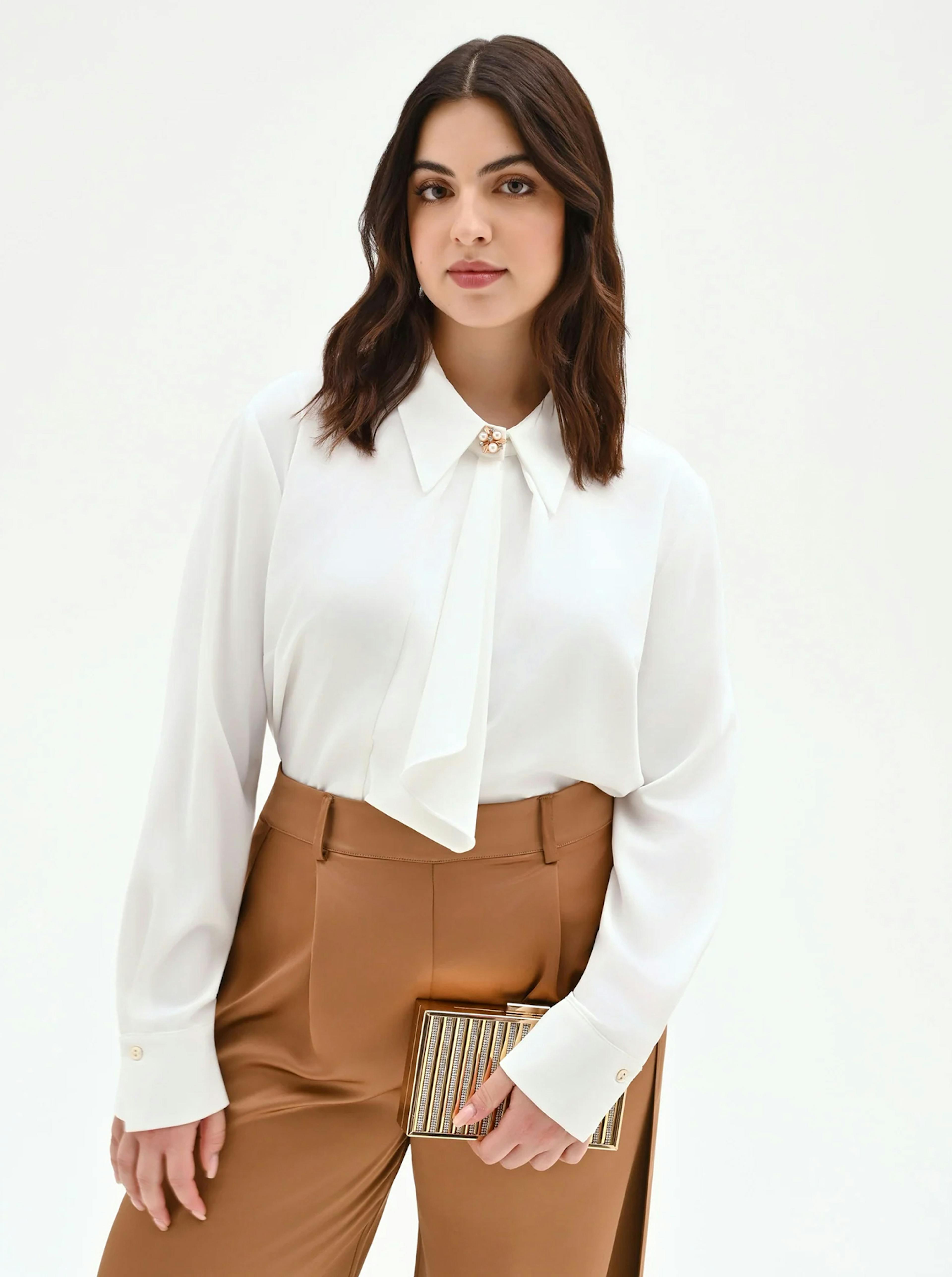 Woman in a white blouse with a bow and brown pants, holding a striped clutch. She has medium-length dark hair and a confident expression. Minimalist background.