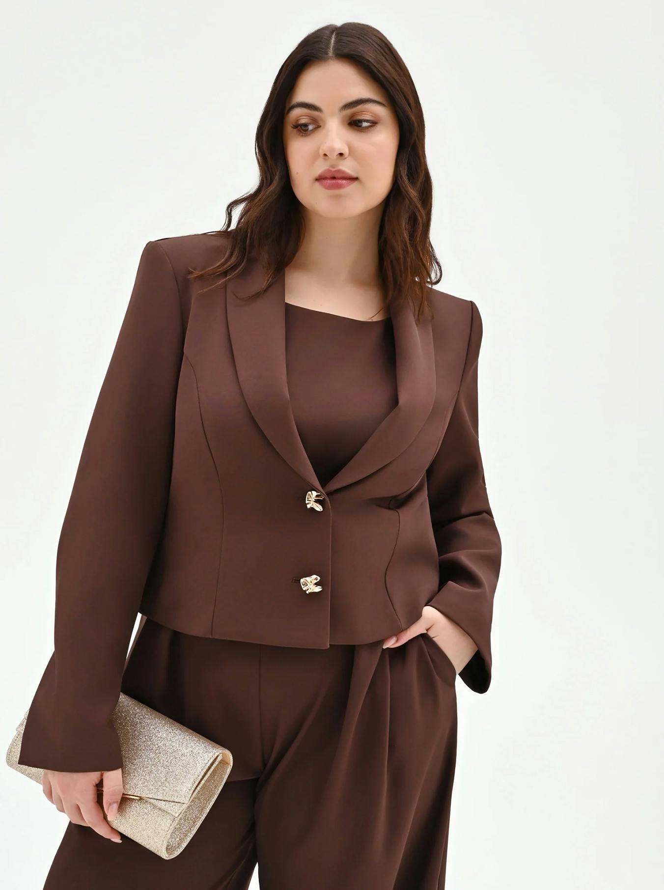 Woman in a stylish brown tailored suit with gold buttons, holding a shimmering clutch. Her expression is serene, exuding confidence and elegance.