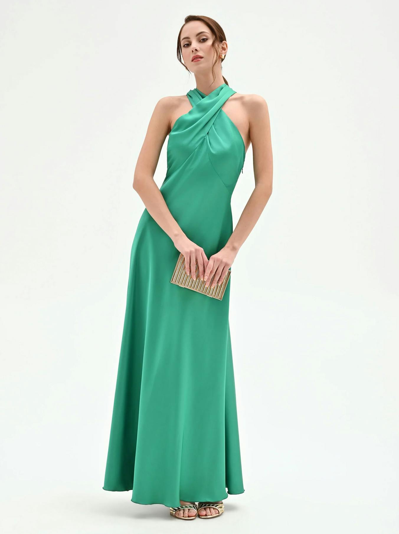 Woman in a stylish green halterneck gown holding a gold clutch. The dress features a long, flowing skirt. She stands poised against a white background.