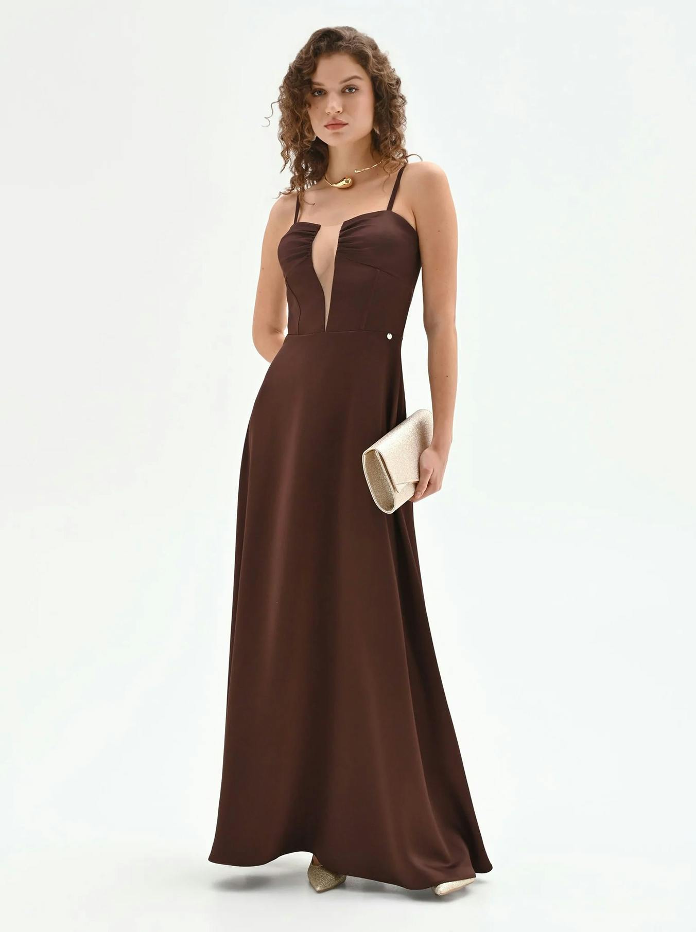Woman in a long brown dress with thin straps and a deep V-neck, holding a beige clutch. She stands against a plain white background, exuding elegance.