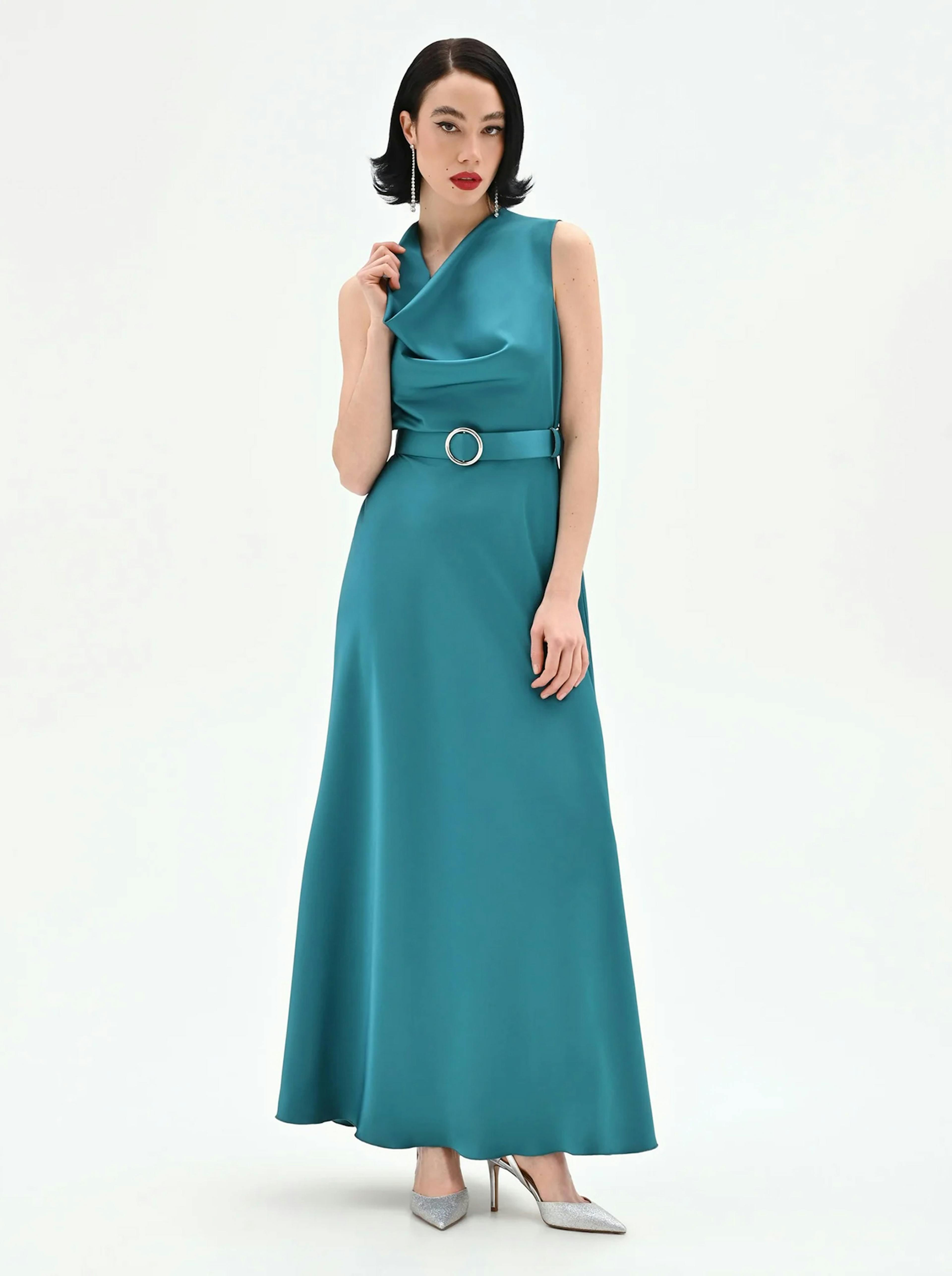 Woman in an elegant teal gown with cowl neckline and belted waist poses against a plain background. She wears silver heels and has sleek, dark hair.