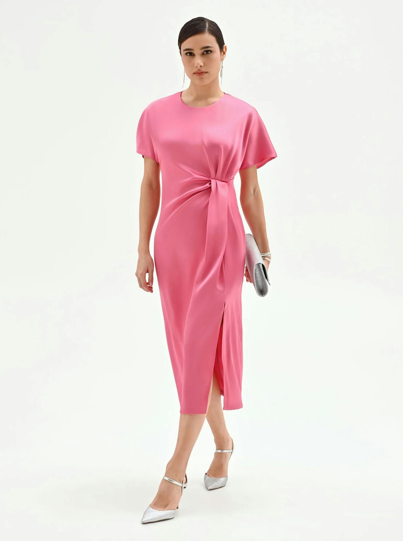 A woman in a sleek, pink dress with a front knot walks forward. She carries a silver clutch and wears matching silver heels. The mood is elegant and poised.