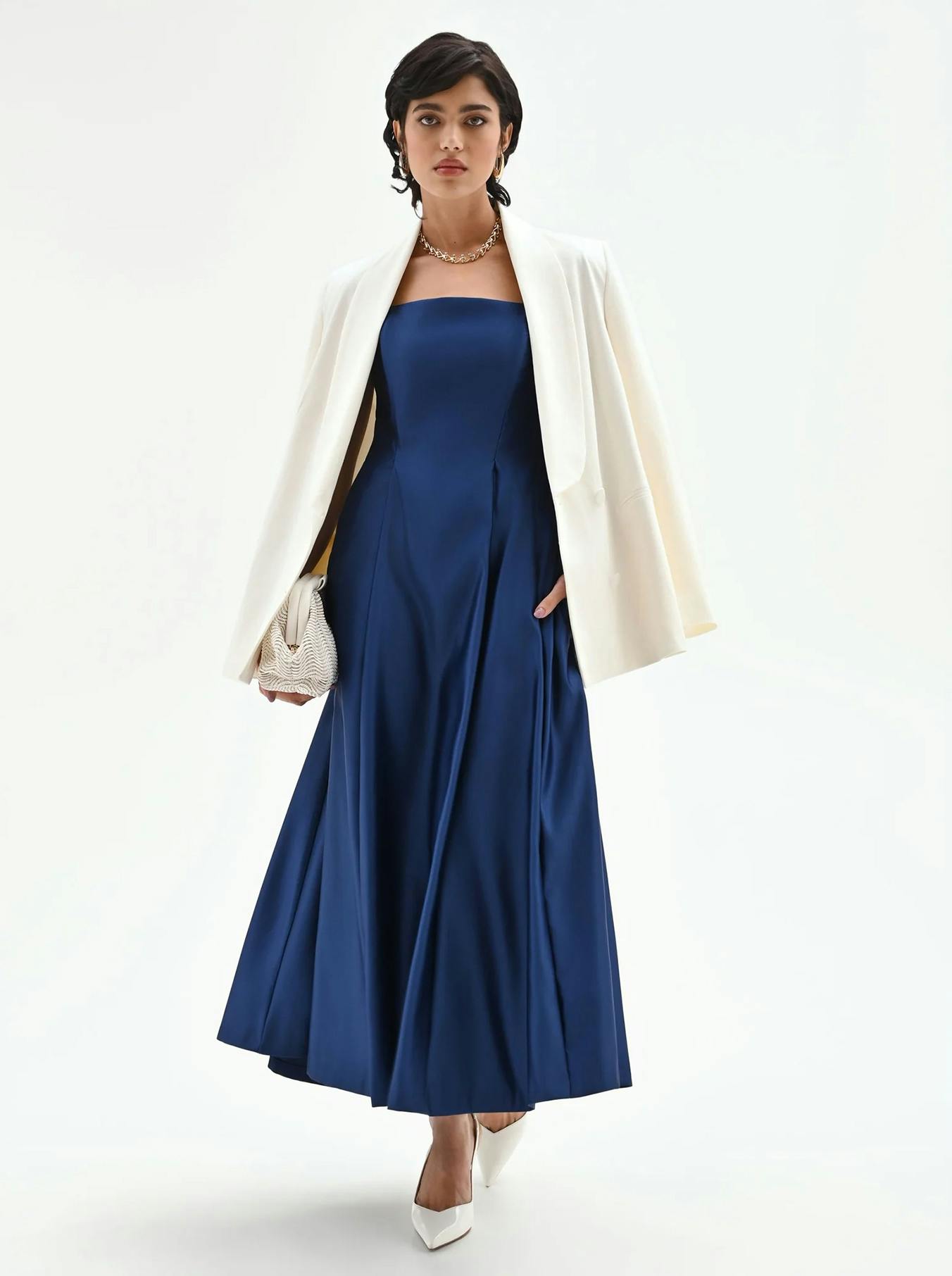 Stylish woman in a blue strapless gown with a white blazer draped over her shoulders. She carries a textured clutch and wears white heels, exuding elegance.