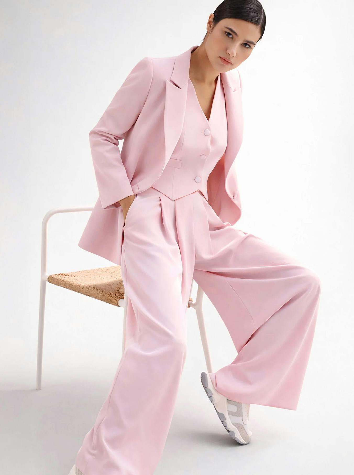A woman sits confidently on a chair wearing a pastel pink suit and sneakers. The mood is stylish and modern, with a white background highlighting the outfit.