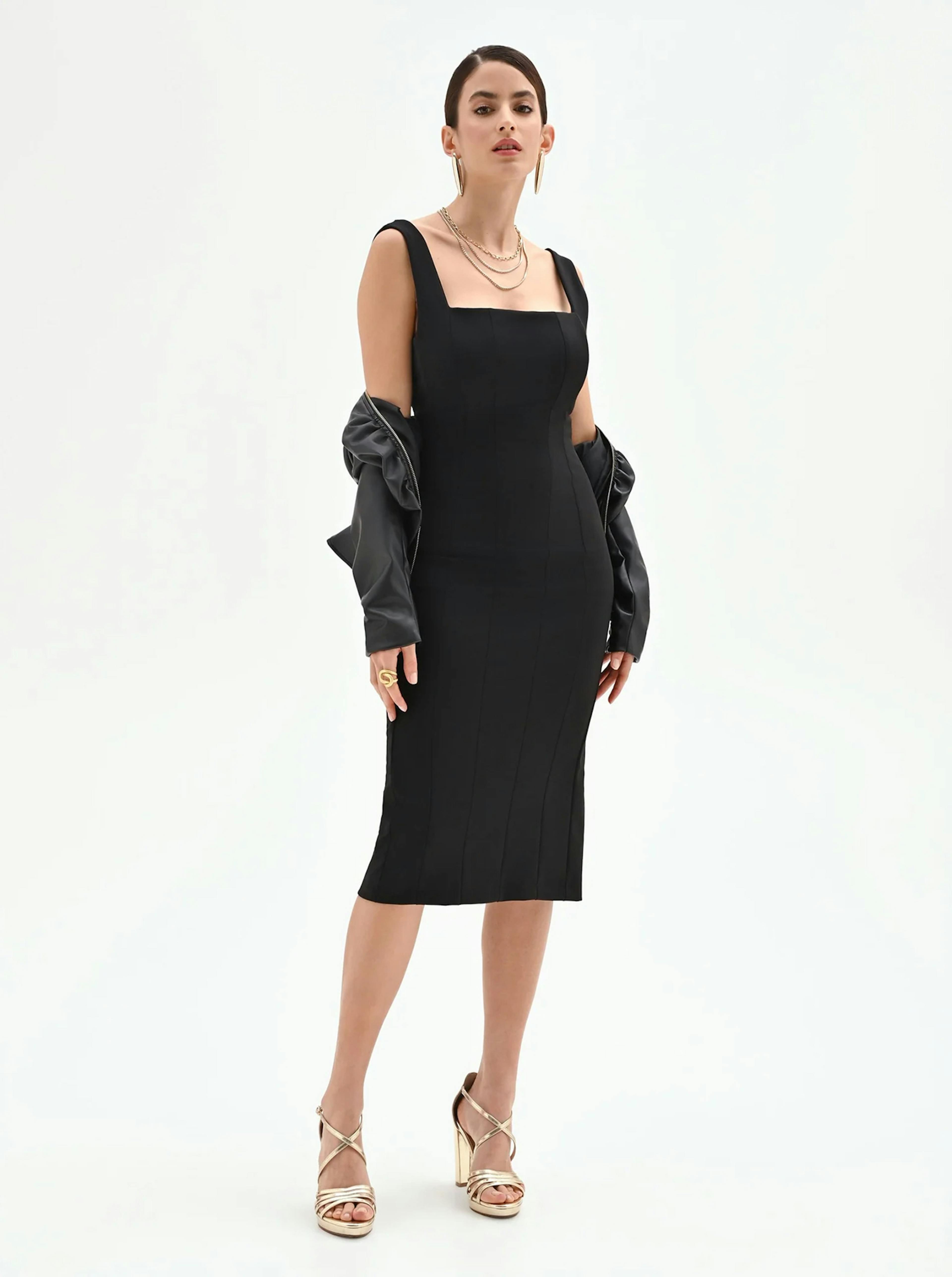 A woman in a sleek black dress with a square neckline stands confidently. She wears gold heels, layered necklaces, a draped leather jacket, and exudes elegance.