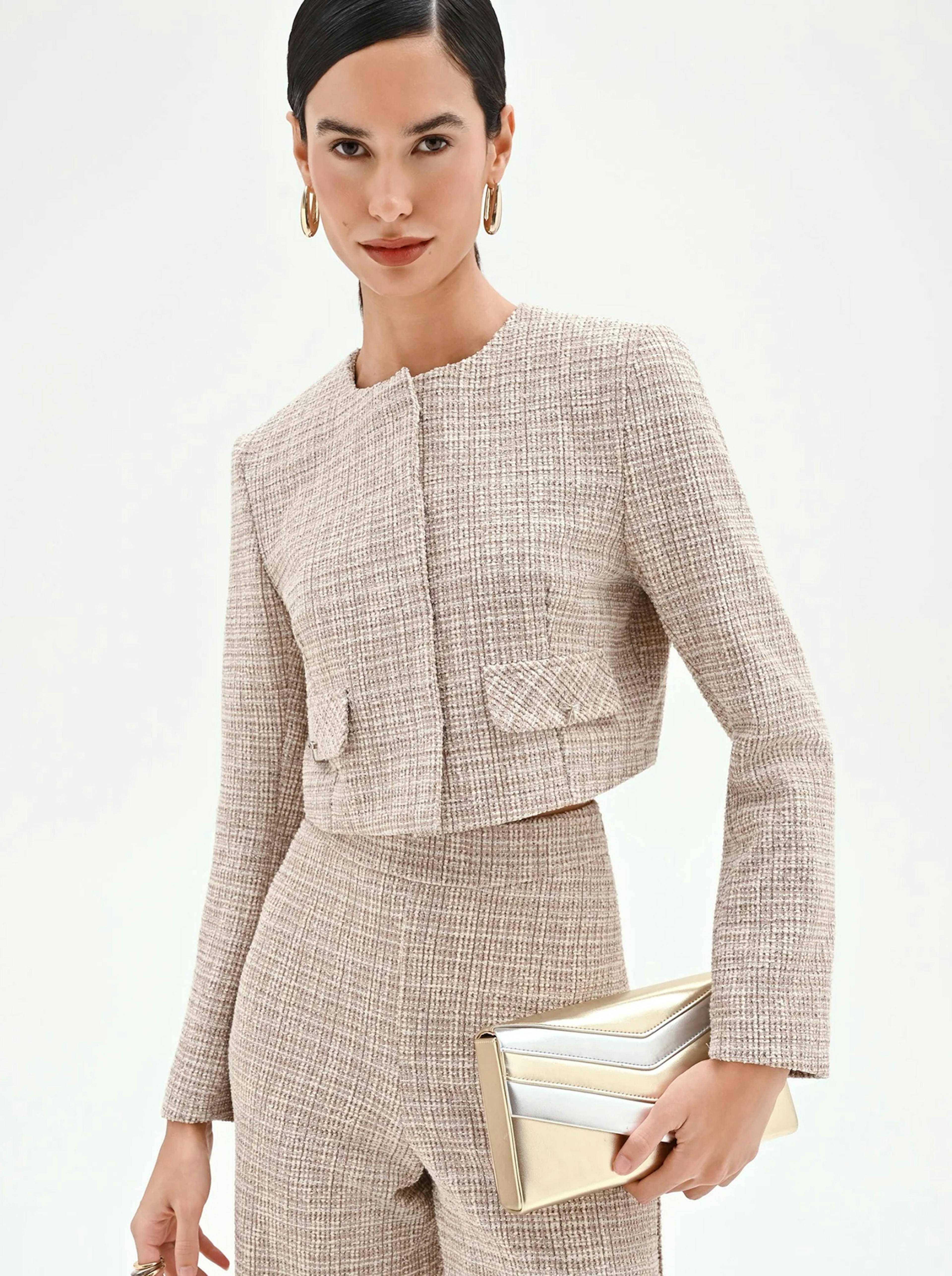 Woman in a beige tweed suit with a cropped jacket and matching high-waisted pants, holding a gold and silver clutch. She wears gold hoop earrings.
