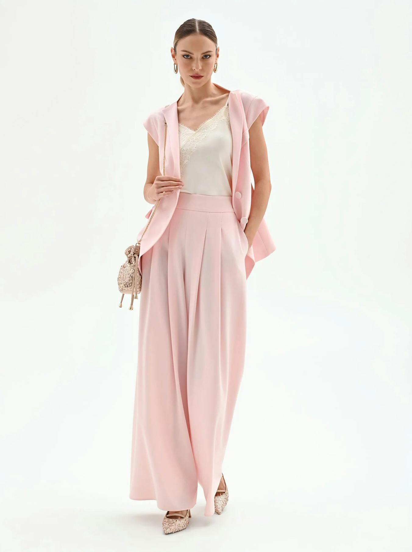 Woman in elegant pink suit with wide-leg trousers, sleeveless blazer, and lace camisole. Holds a small textured handbag, conveying a chic and confident style.