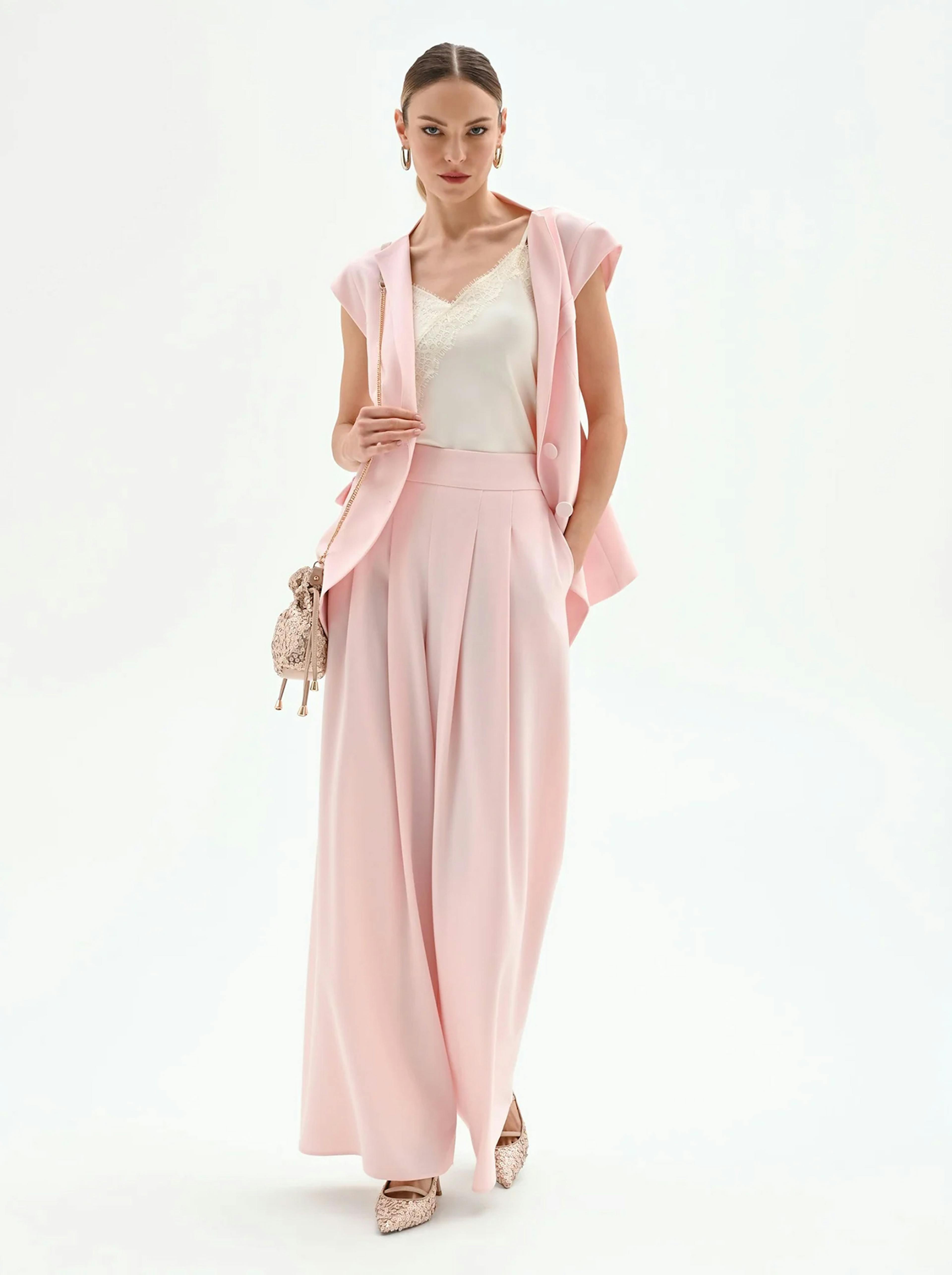 Woman in elegant pink suit with wide-leg trousers, sleeveless blazer, and lace camisole. Holds a small textured handbag, conveying a chic and confident style.