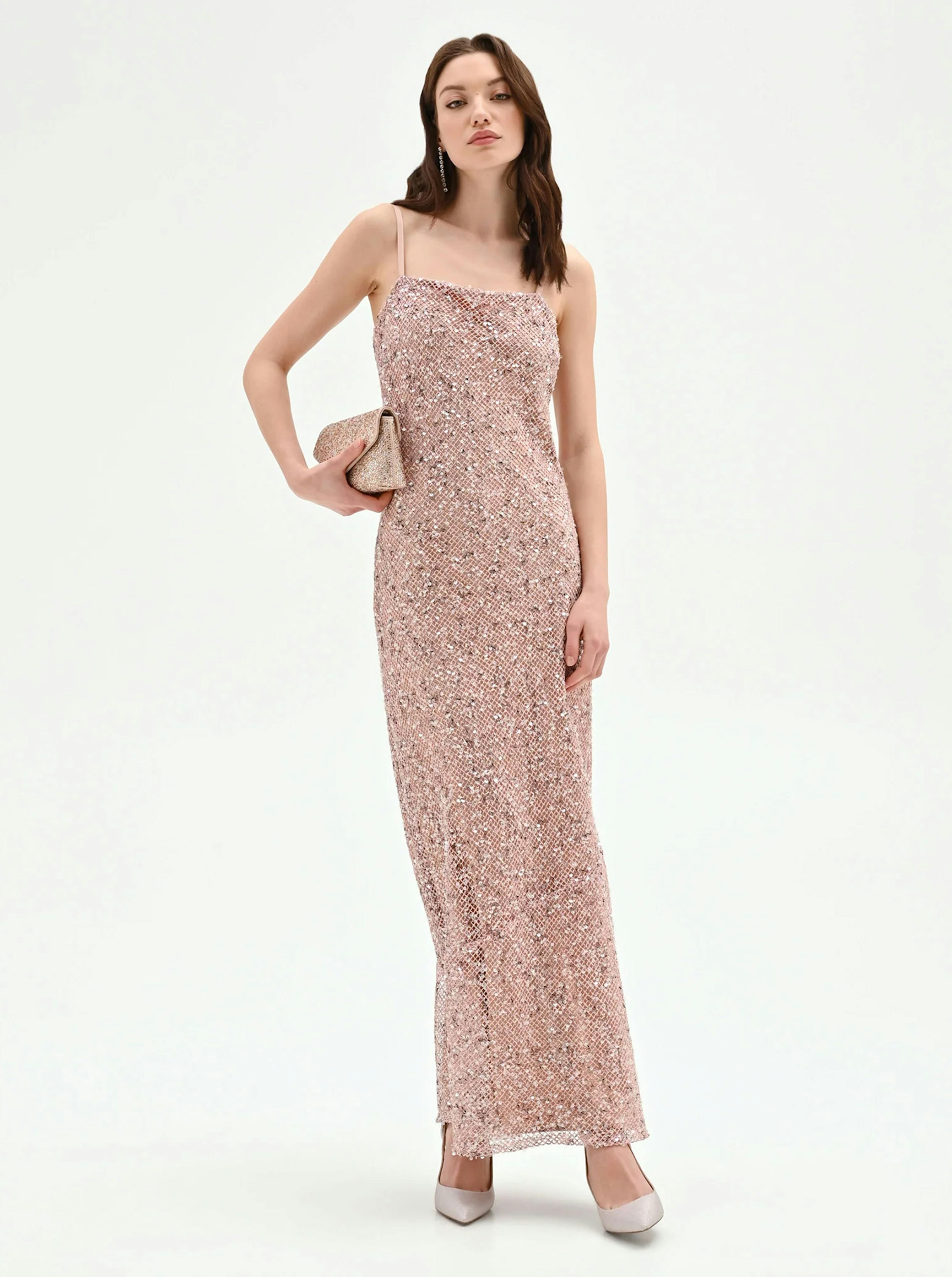 Woman in a sparkling pink sequin gown holds a matching clutch. She stands confidently against a white background, exuding elegance and poise.