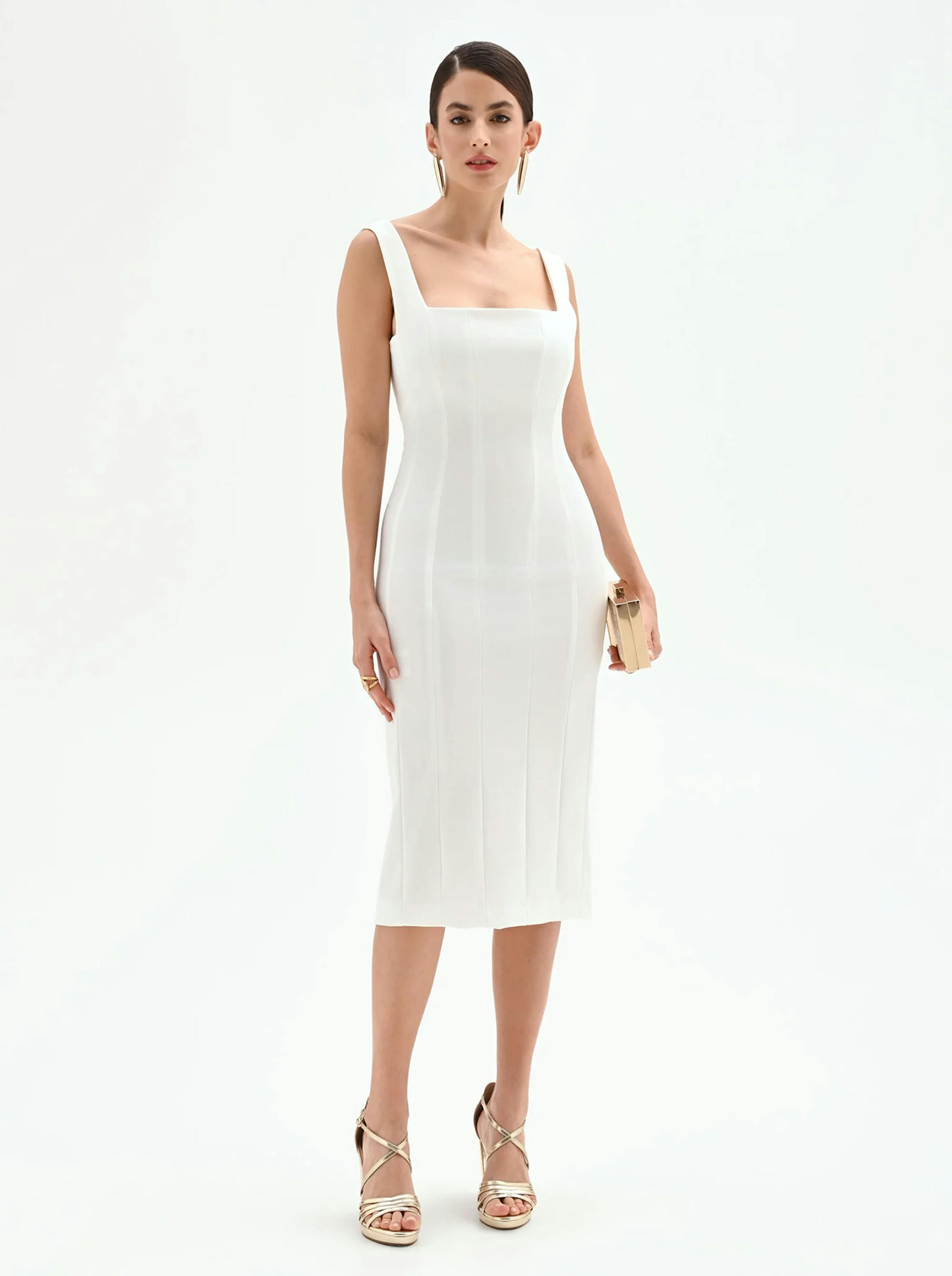 Woman in a sleek, sleeveless white midi dress and gold heels stands confidently holding a small clutch. Minimalistic, elegant, and poised.