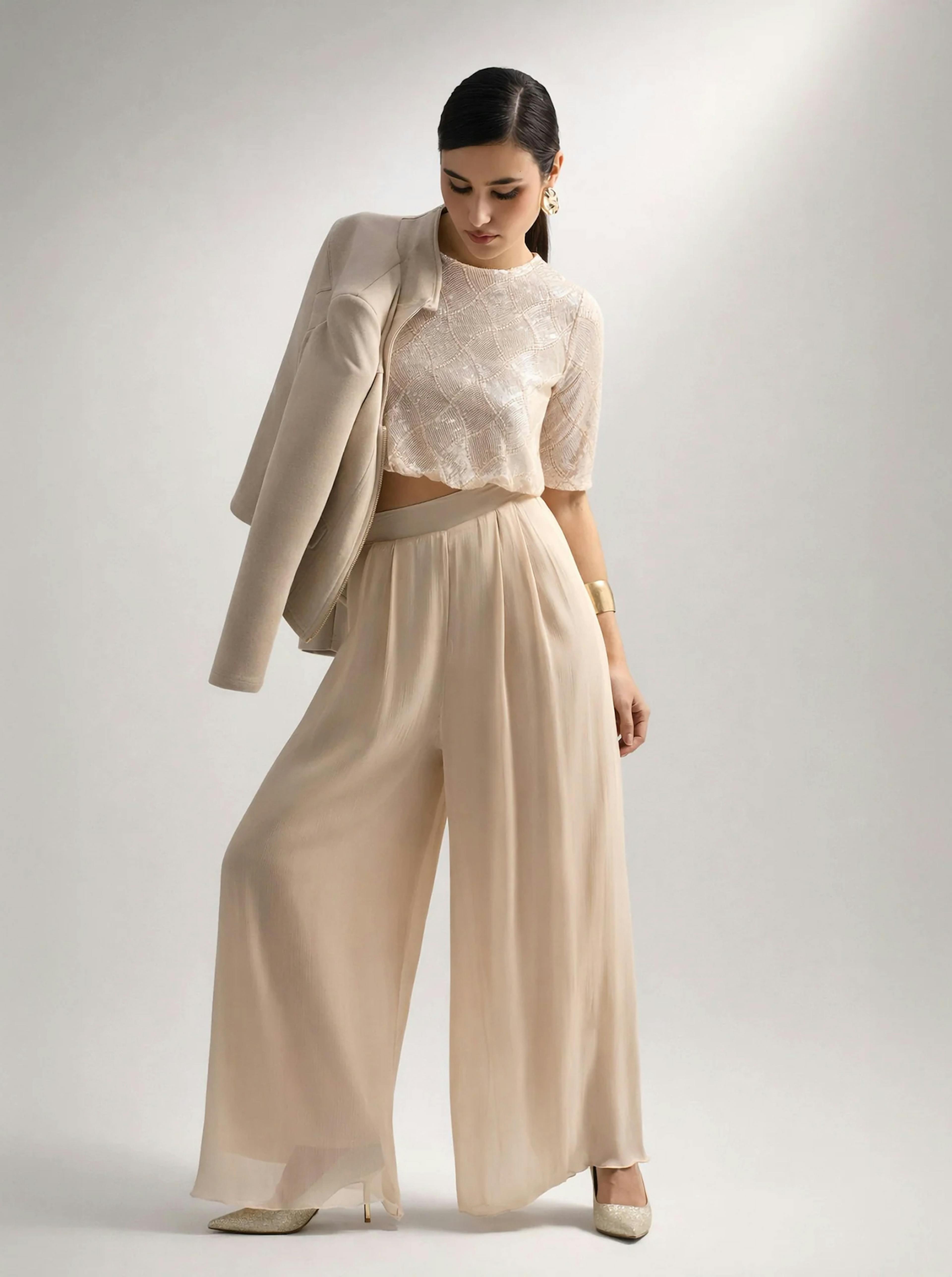 A woman in an elegant, neutral-toned outfit poses confidently. She wears a lace crop top, wide-leg trousers, and a draped blazer, exuding sophistication.