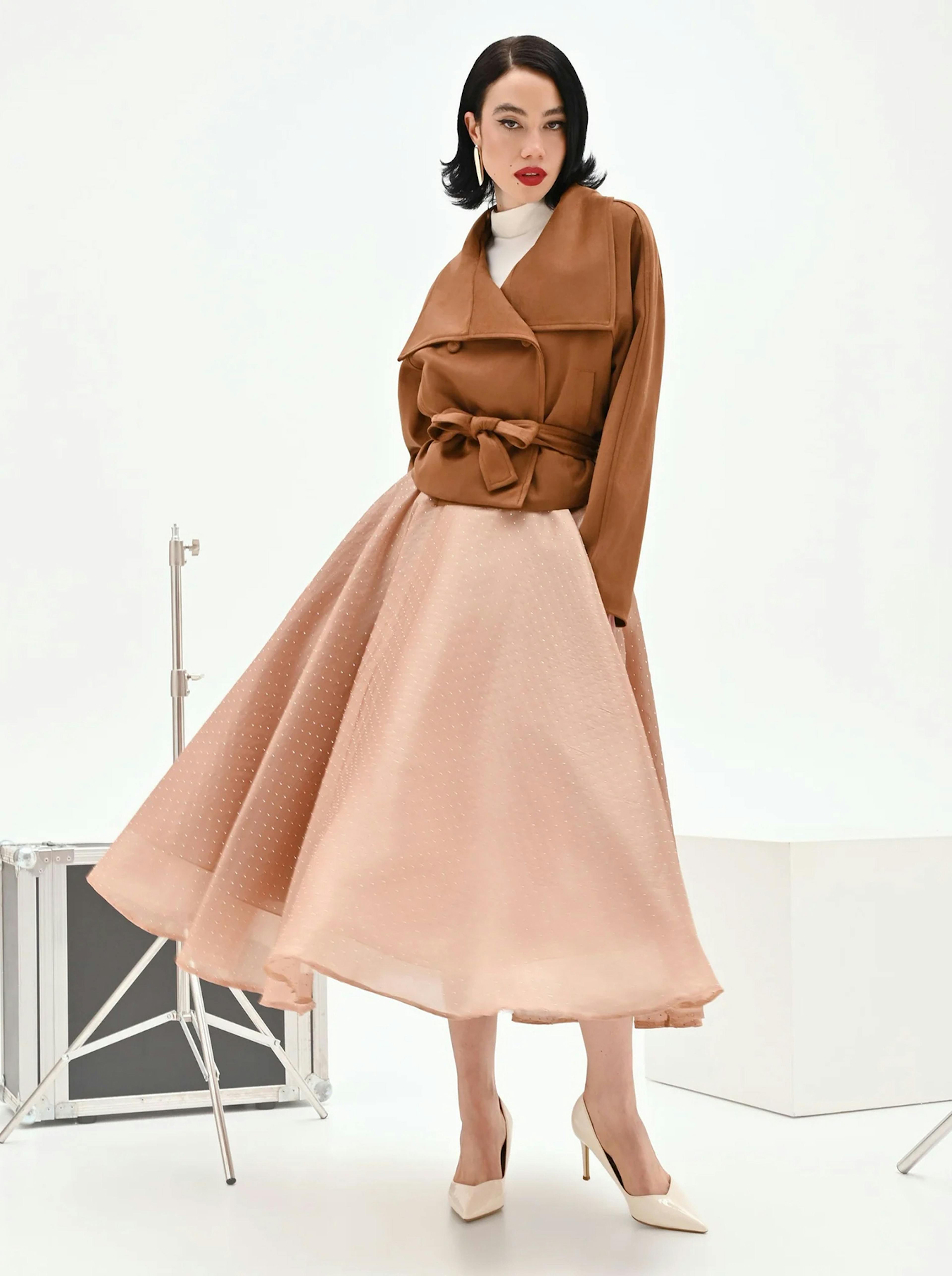 Sophisticated woman in brown jacket and flowing pink skirt, posing elegantly in a studio. Her expression is confident, exuding vintage charm.