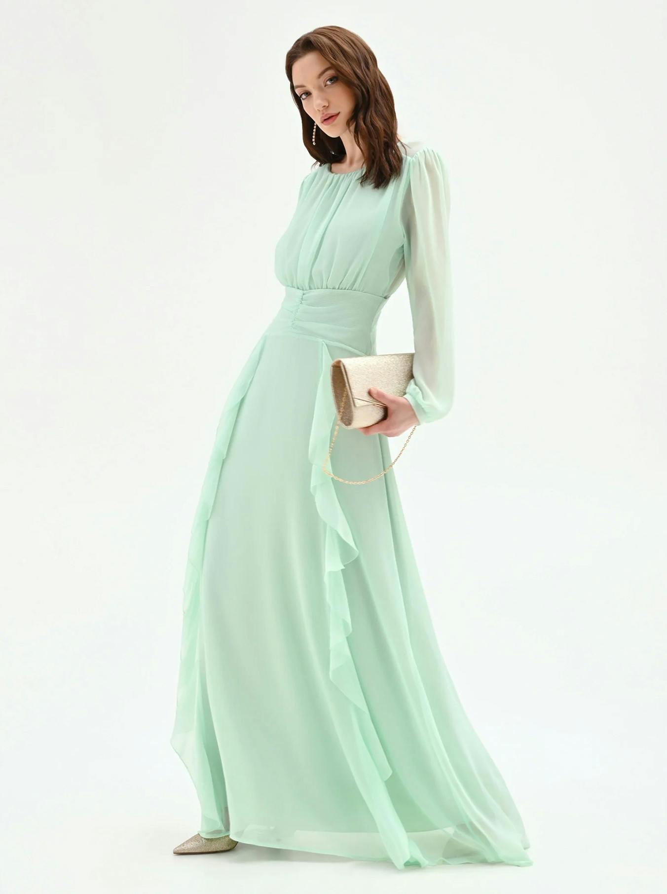 A person stands in a flowing mint green dress with long sleeves and ruffled detailing, holding a gold clutch.