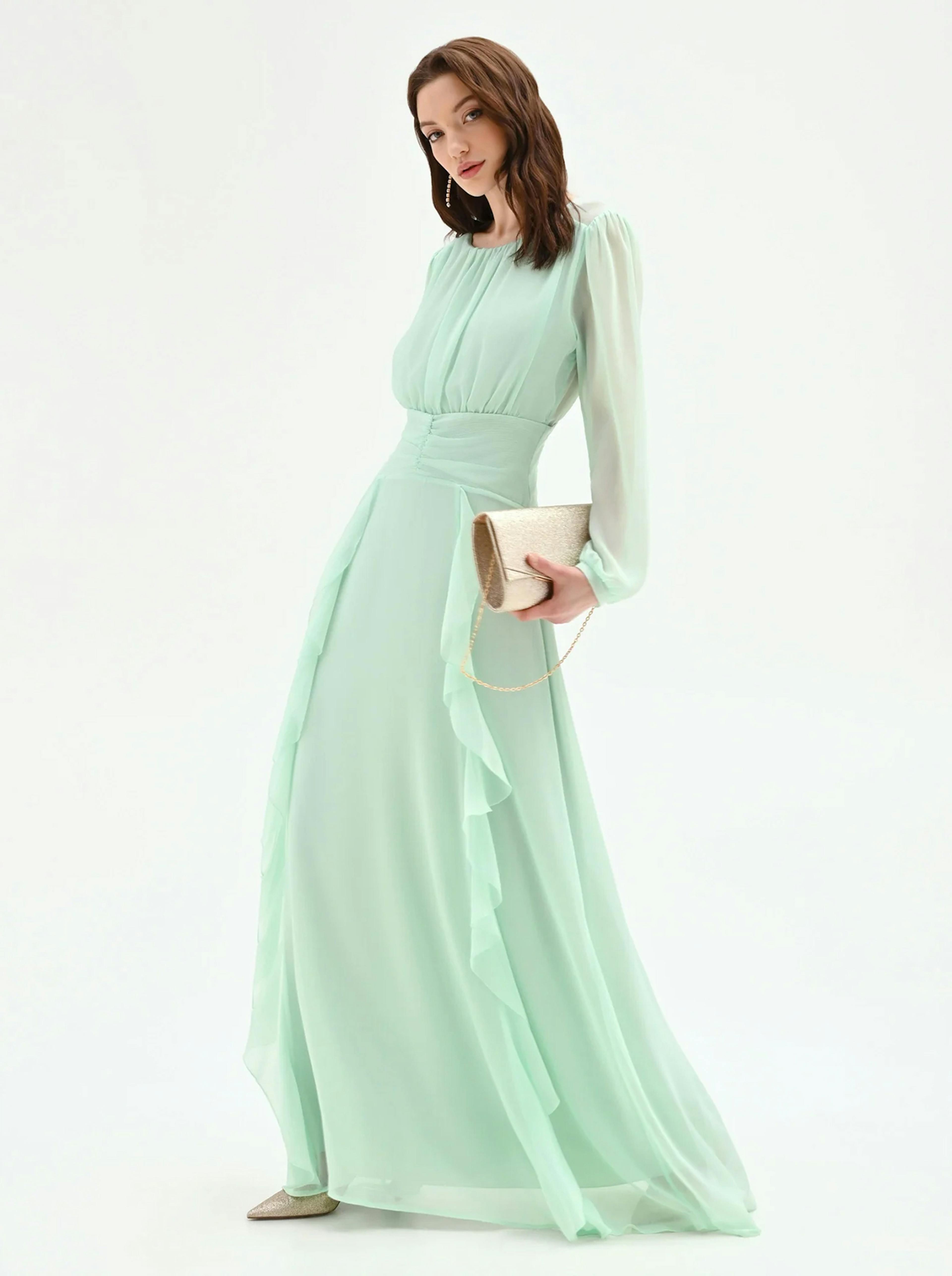 A person stands in a flowing mint green dress with long sleeves and ruffled detailing, holding a gold clutch.