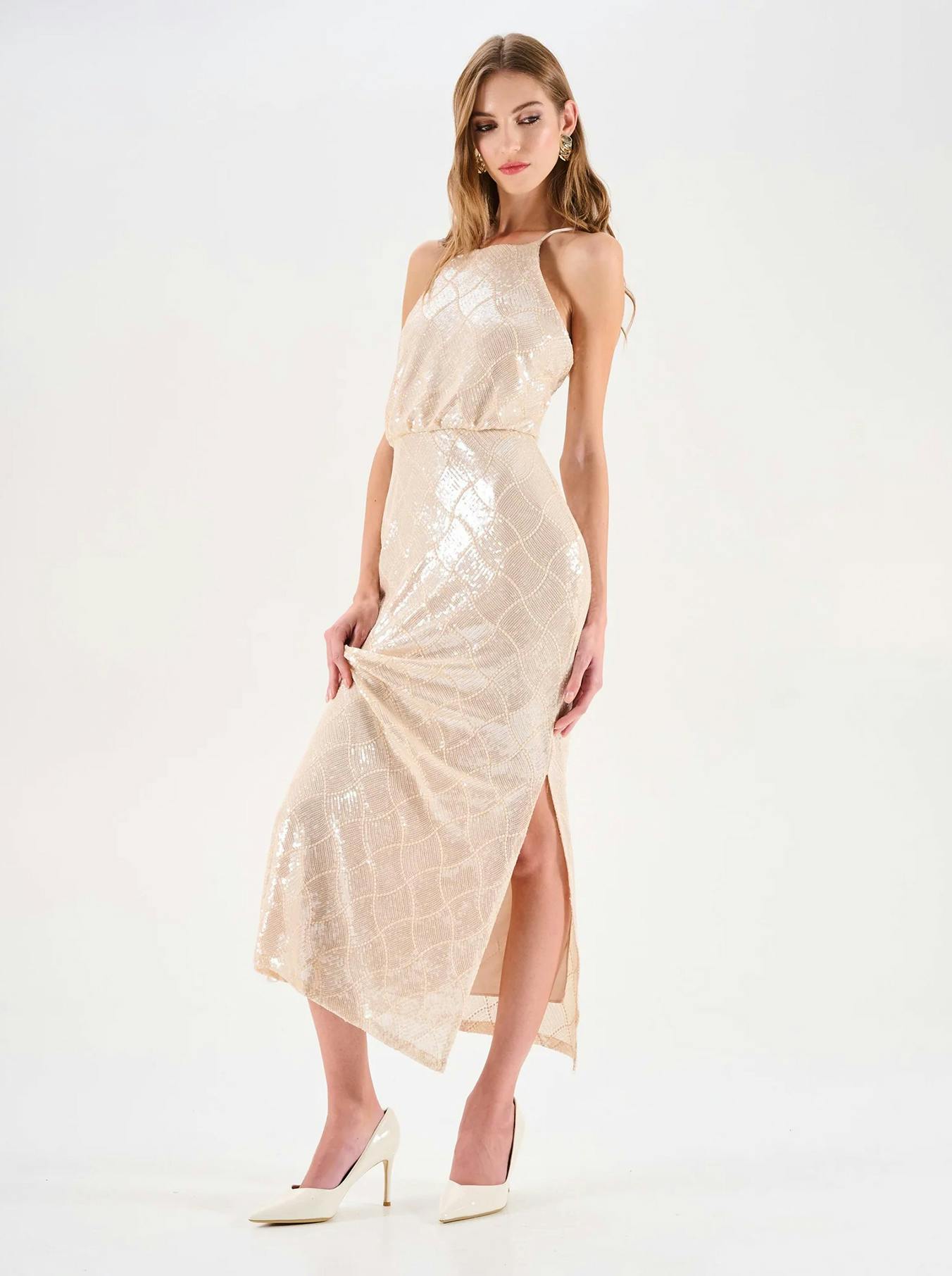 A model poses in a shimmering beige maxi dress with a thigh-high slit, complemented by white high heels against a clean backdrop.