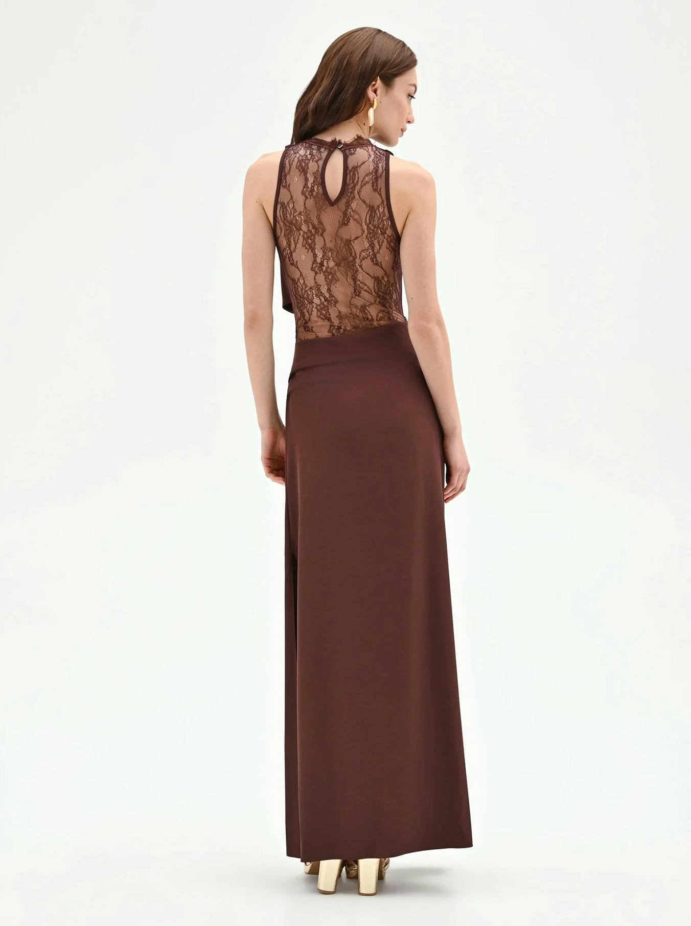 A woman stands with her back turned, showcasing a brown gown with a lace bodice and a keyhole detail, paired with elegant gold heels.