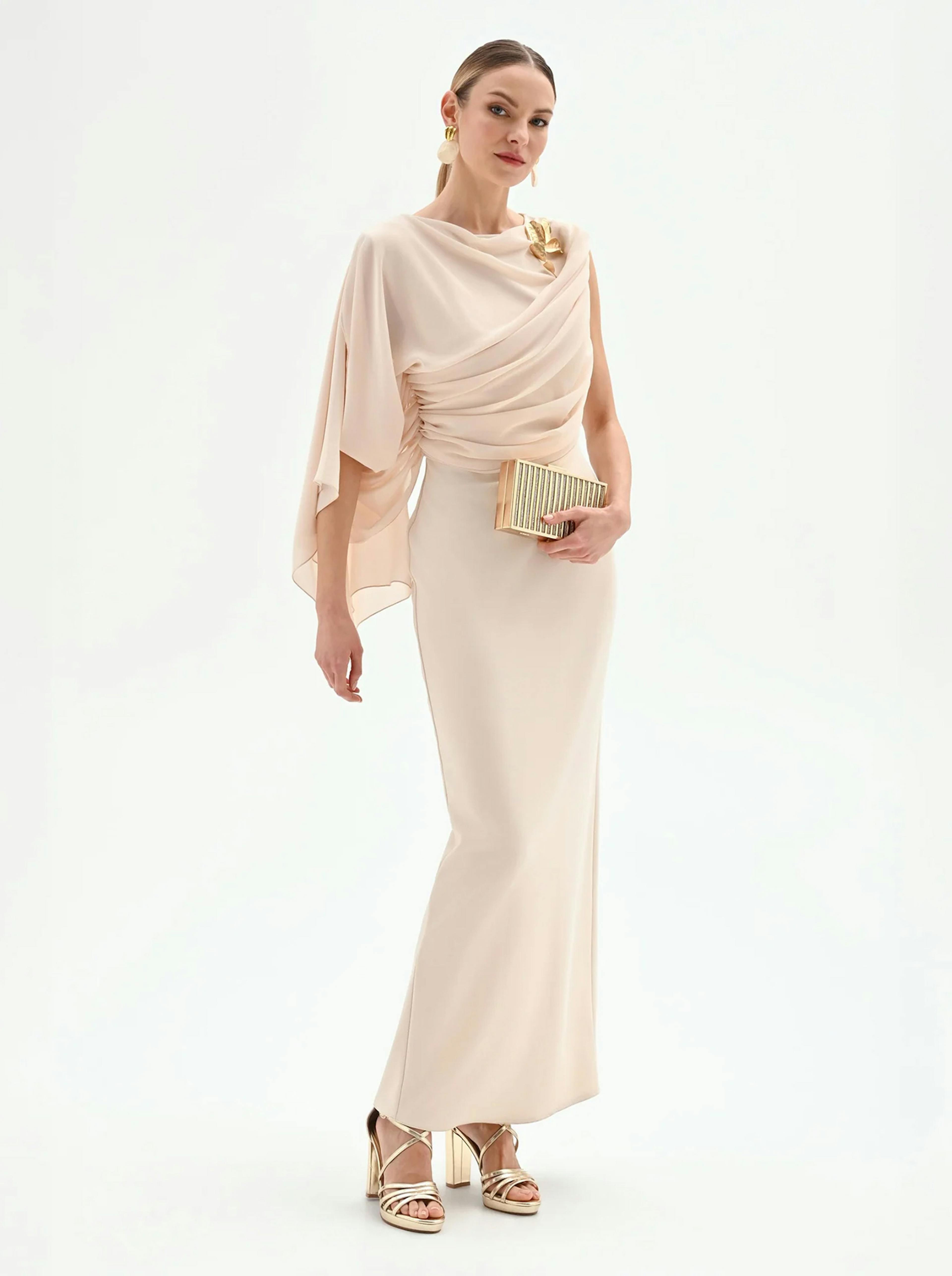 Model in a fitted, elegant beige gown with draped detailing, holding a small clutch, and wearing stylish heels.
