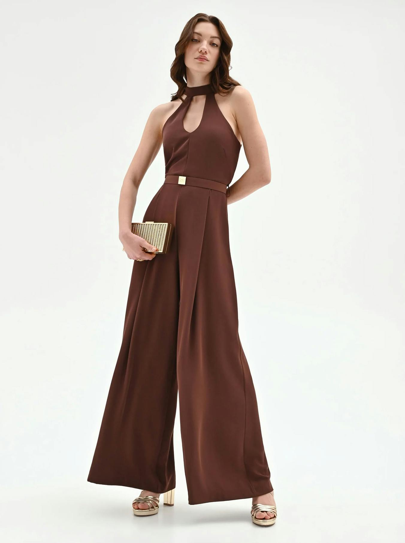 A woman poses in a stylish brown jumpsuit with a halter neckline, holding a gold clutch and wearing metallic platform sandals.