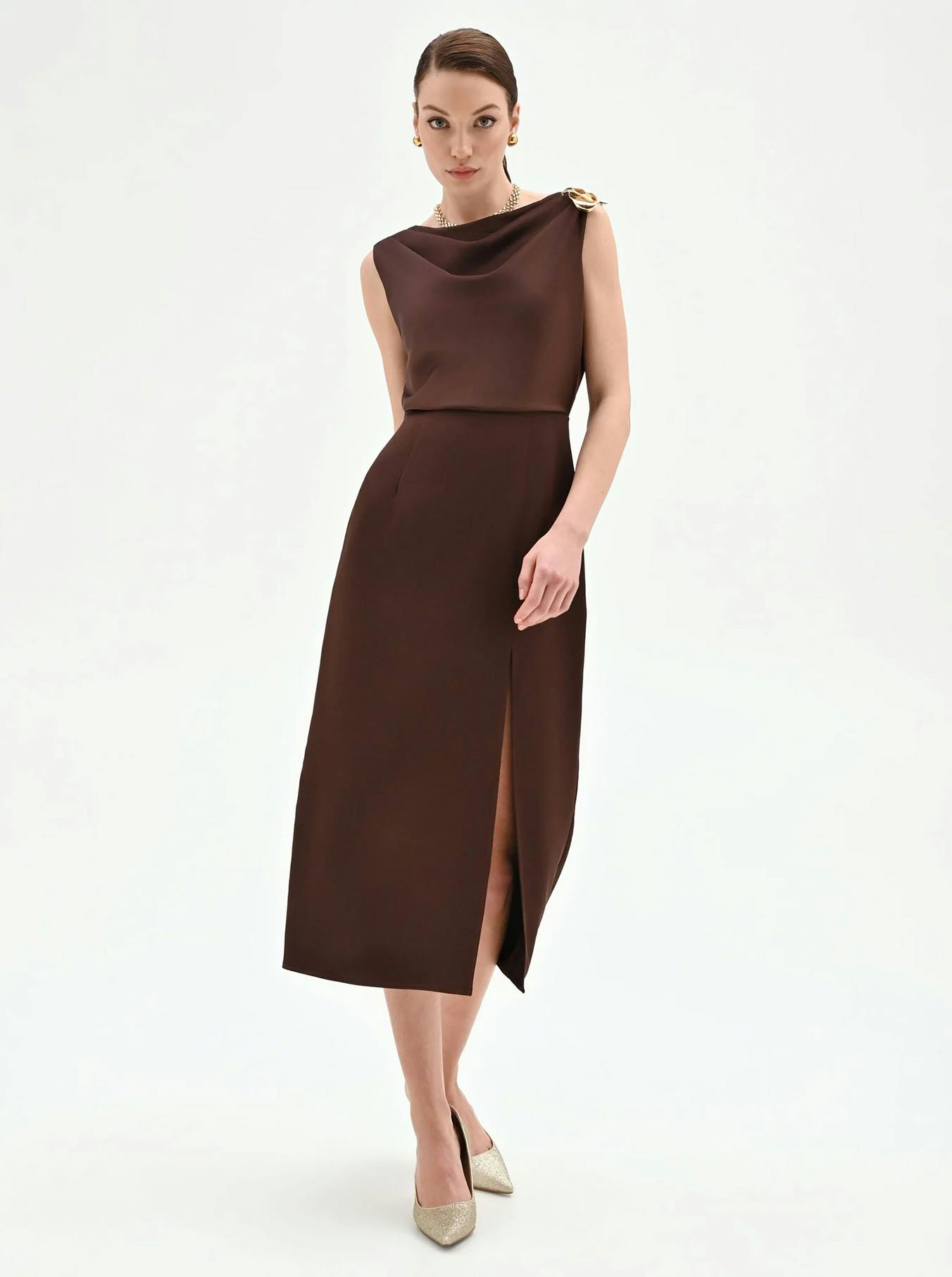 A model showcases a sleek, brown, sleeveless dress featuring a stylish draped neckline and a thigh-high slit, paired with elegant heels.