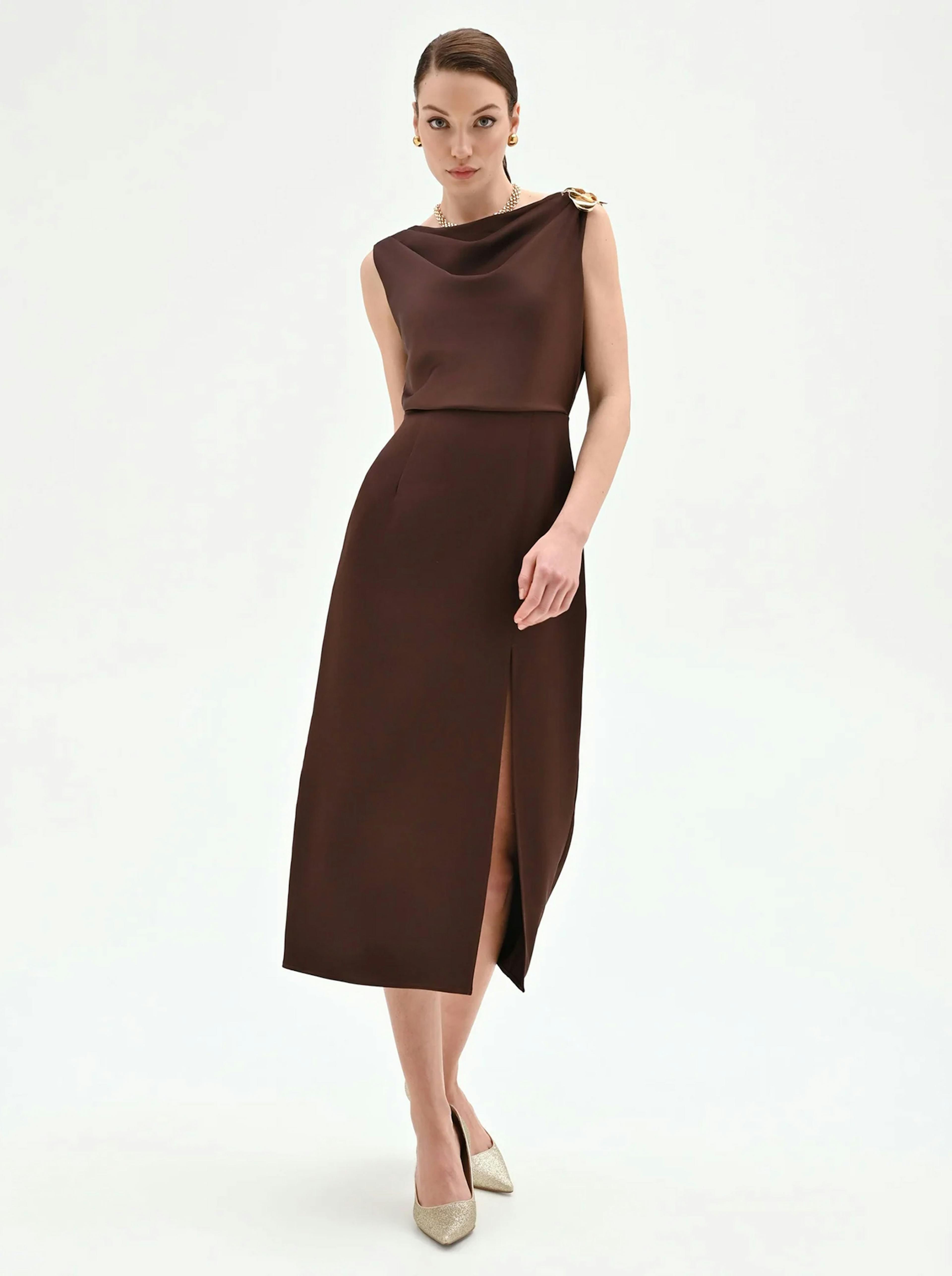 A model showcases a sleek, brown, sleeveless dress featuring a stylish draped neckline and a thigh-high slit, paired with elegant heels.
