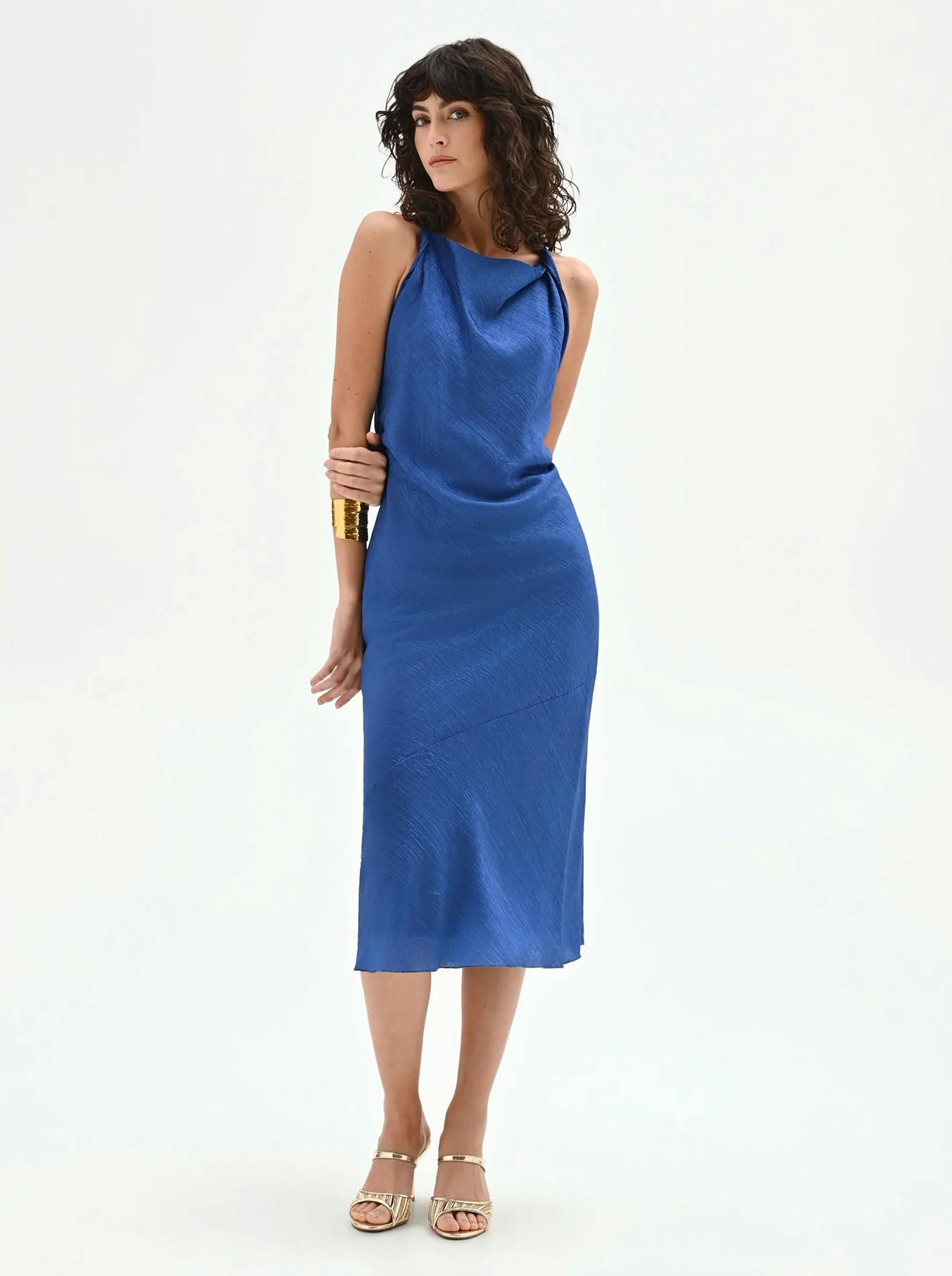 Royal blue midi slip dress in a shimmering textured satin fabric. Featuring a sophisticated draped cowl neckline and a sleeveless silhouette, styled with gold cuff jewelry and metallic sandals.
