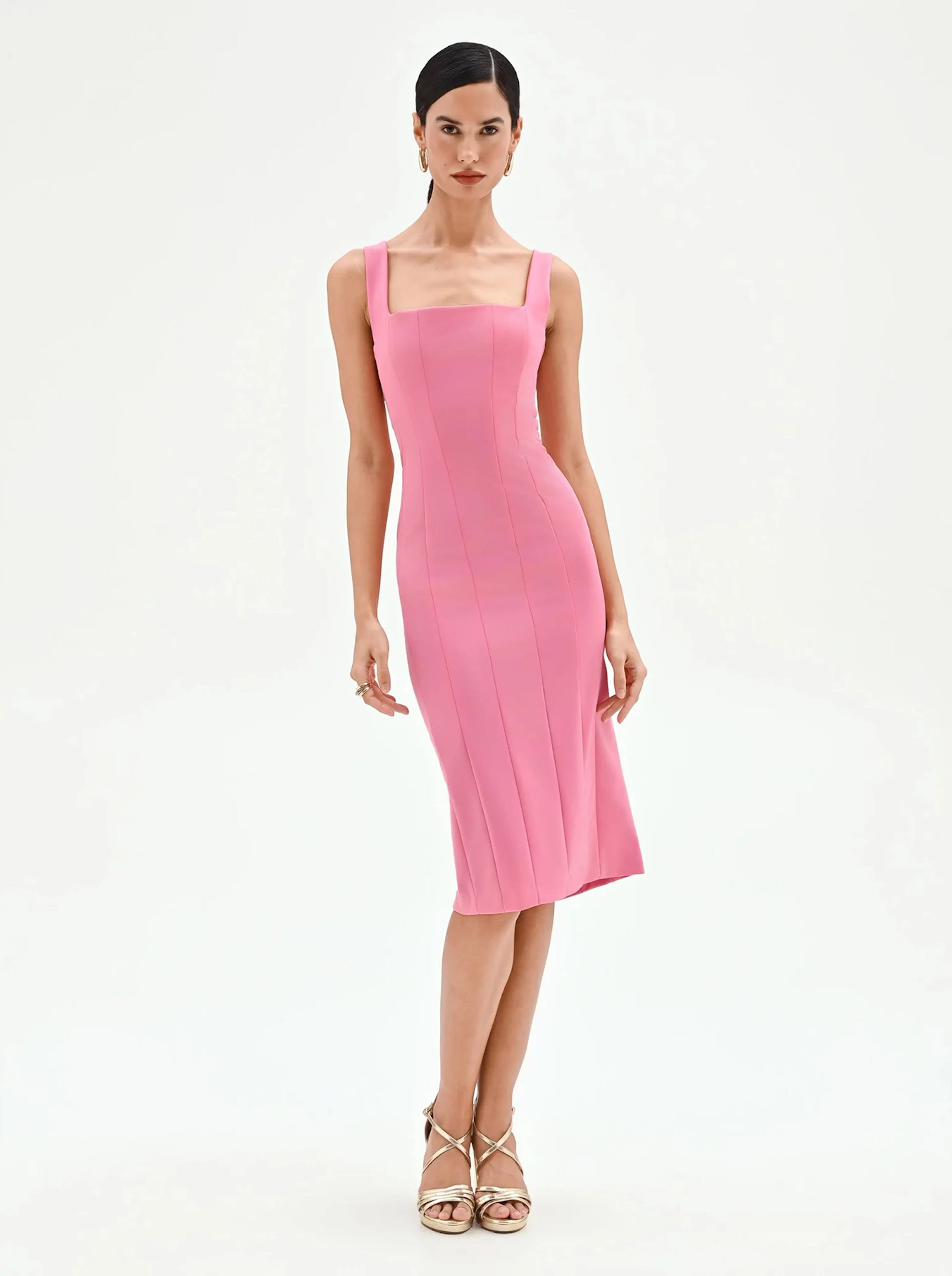 Woman wearing a sleeveless, fitted pink midi dress with square neckline and gold strappy high-heeled sandals, standing against a white background.