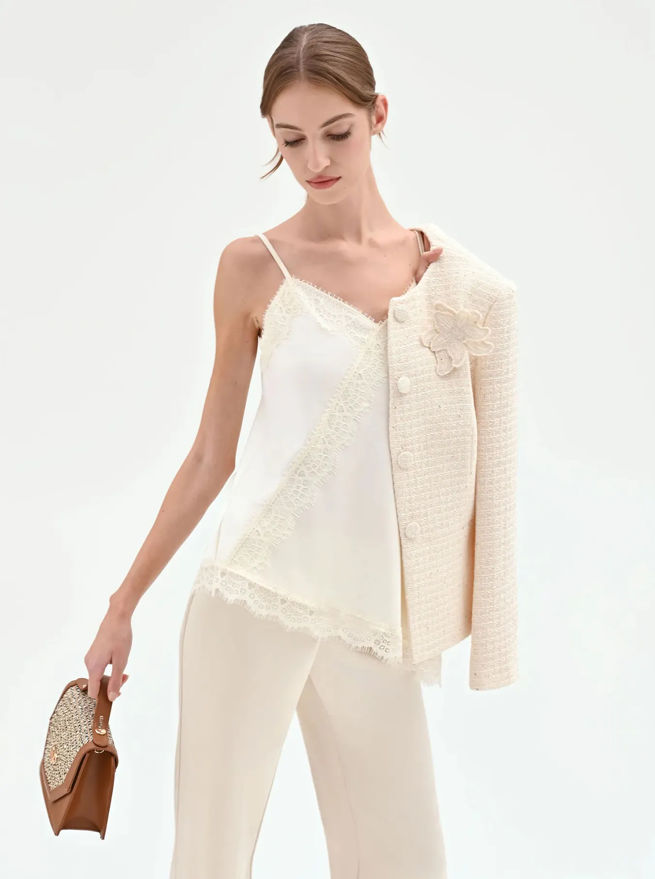Woman in cream lace-trimmed camisole and pants, holding a beige textured jacket over one shoulder and a brown patterned handbag.
