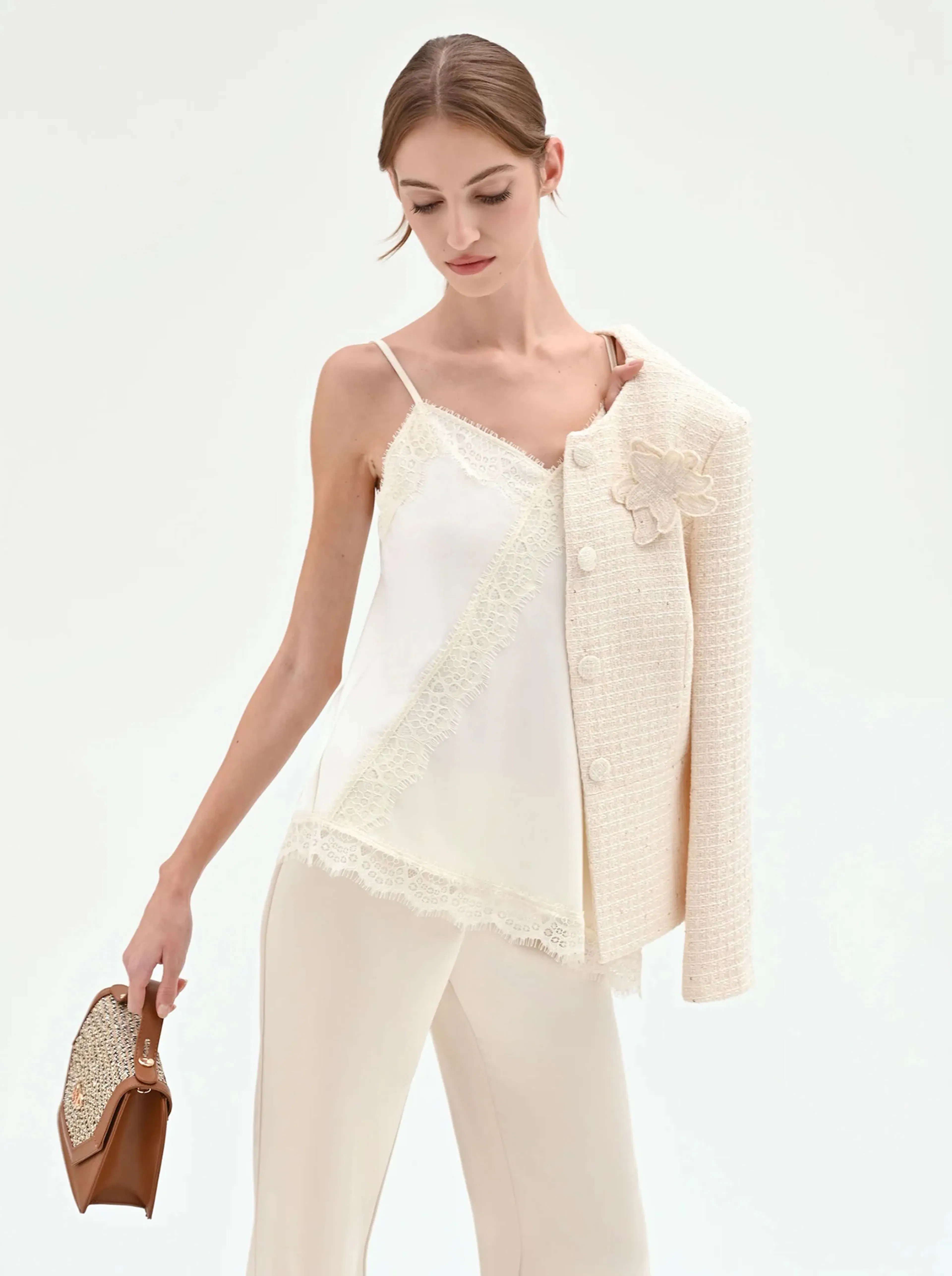 Woman in cream lace-trimmed camisole and pants, holding a beige textured jacket over one shoulder and a brown patterned handbag.