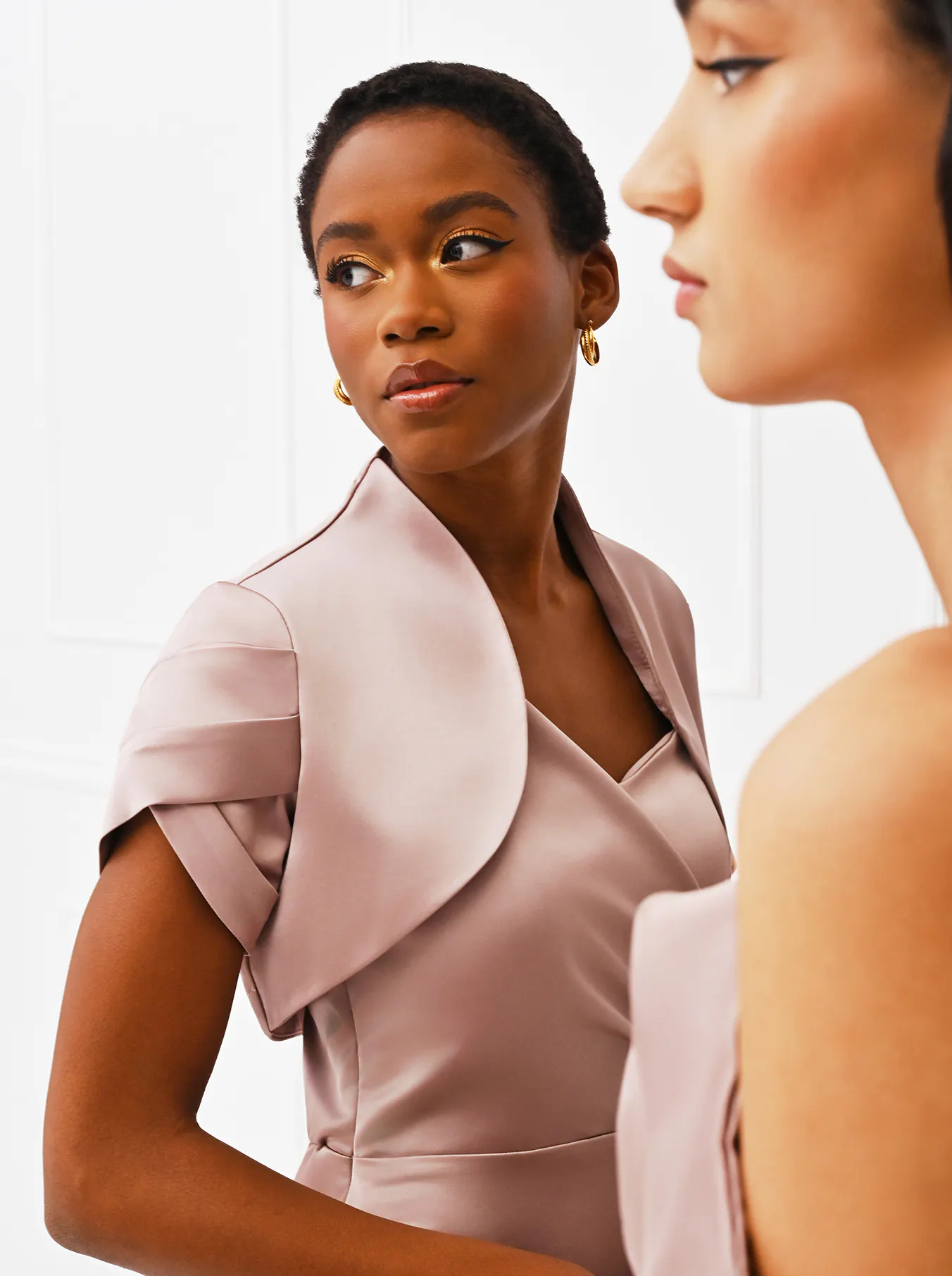 Two women in elegant satin dresses are seen in a close-up. One wears a dress with a shawl collar and the other has an off-the-shoulder design.