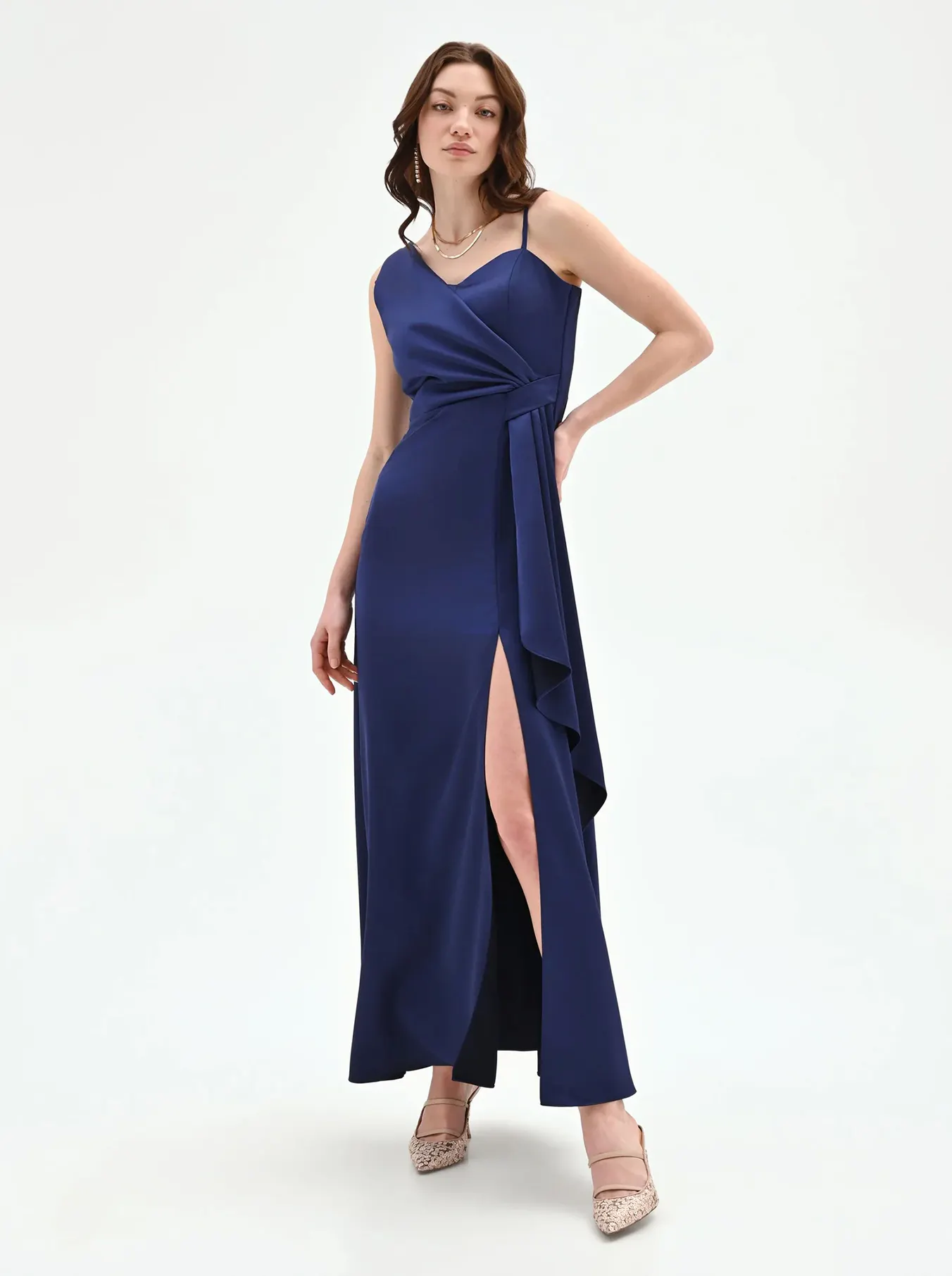 A model poses in a sleek navy gown with a deep side slit and flowing fabric, paired with stylish shoes. The background is plain white.