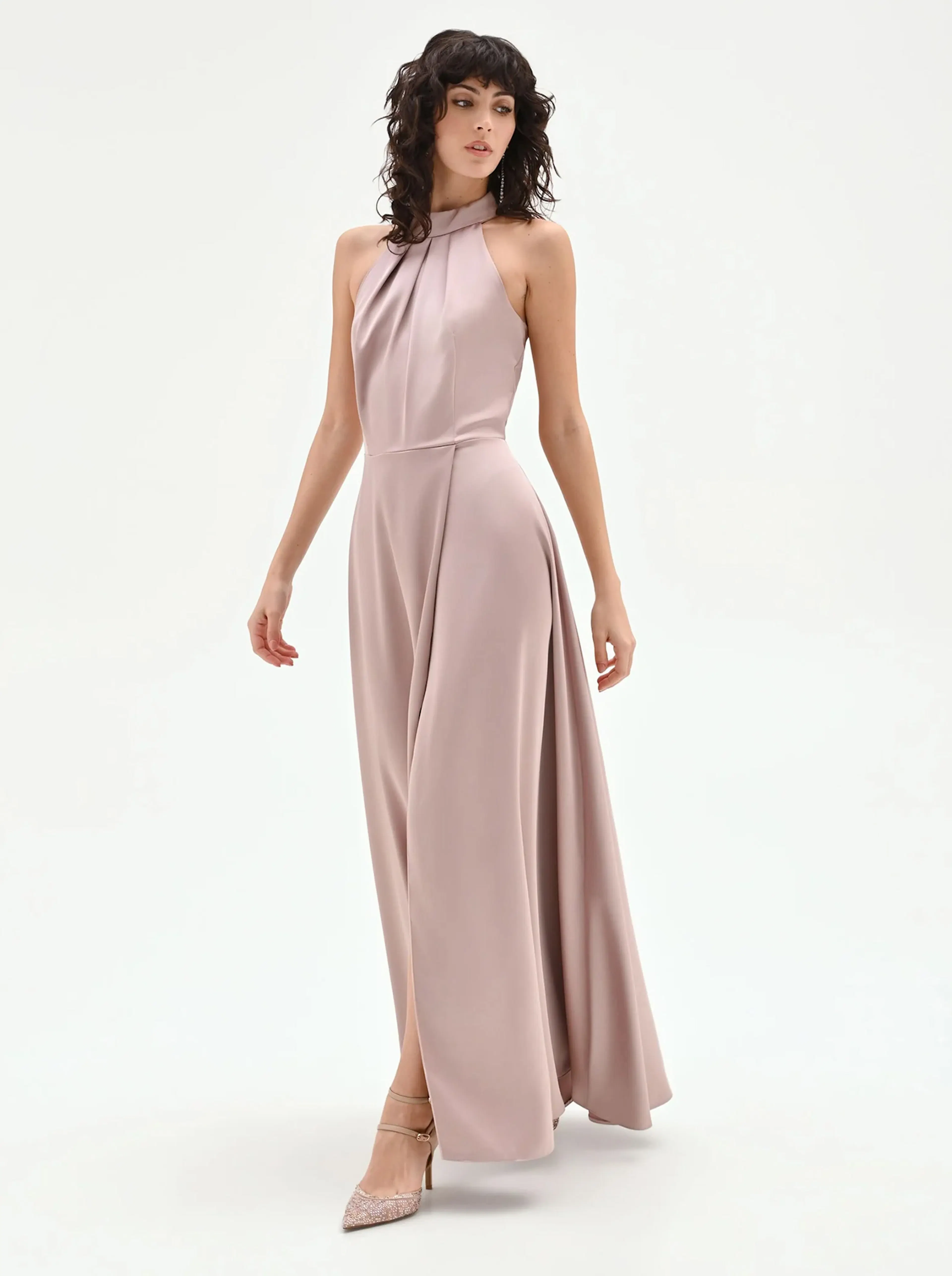 A model in a long, flowy blush gown with a halter neckline, paired with embellished heels, poses against a neutral background.