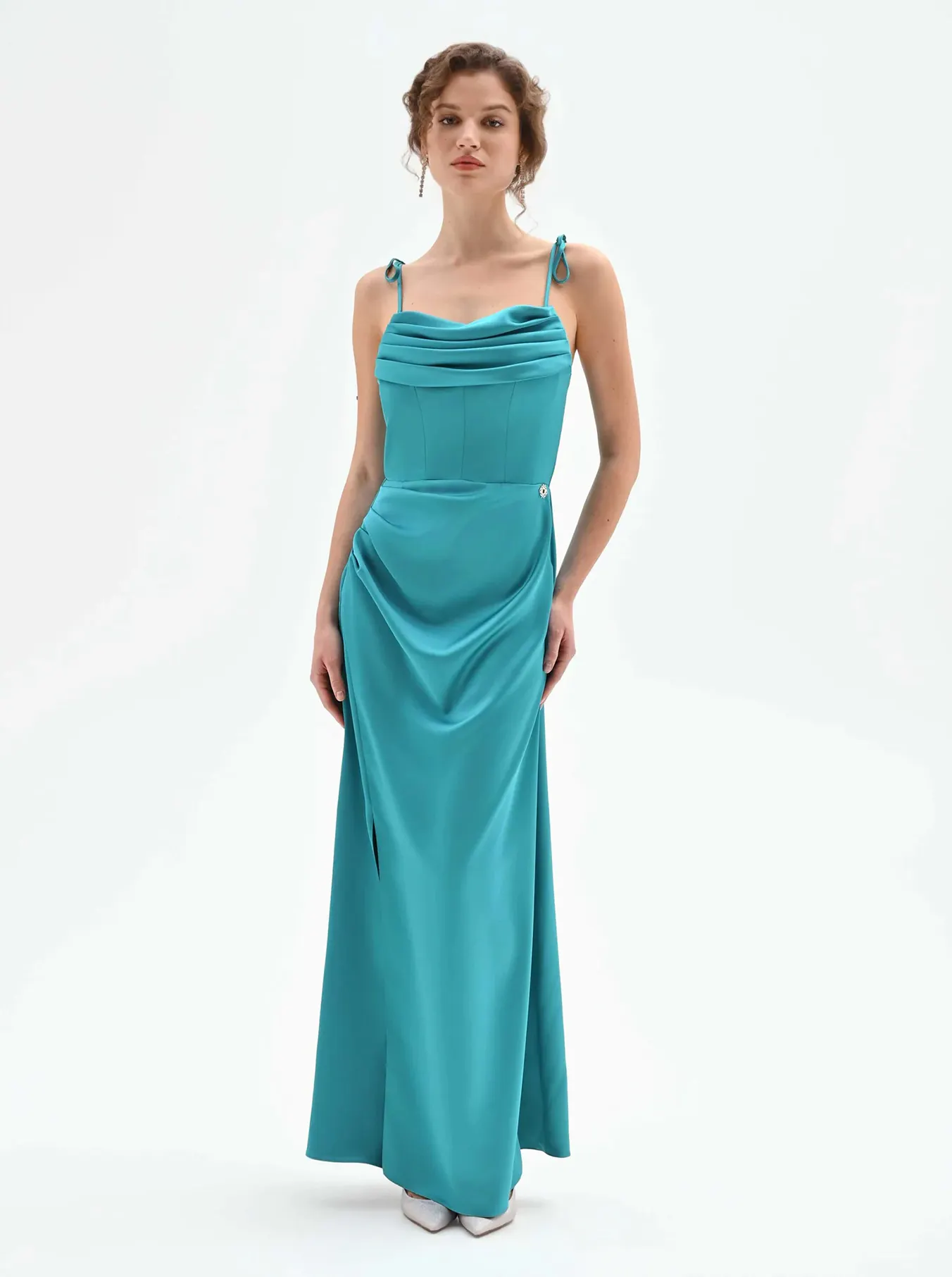 A person wearing a teal, floor-length gown with thin shoulder straps stands facing forward against a plain white background.