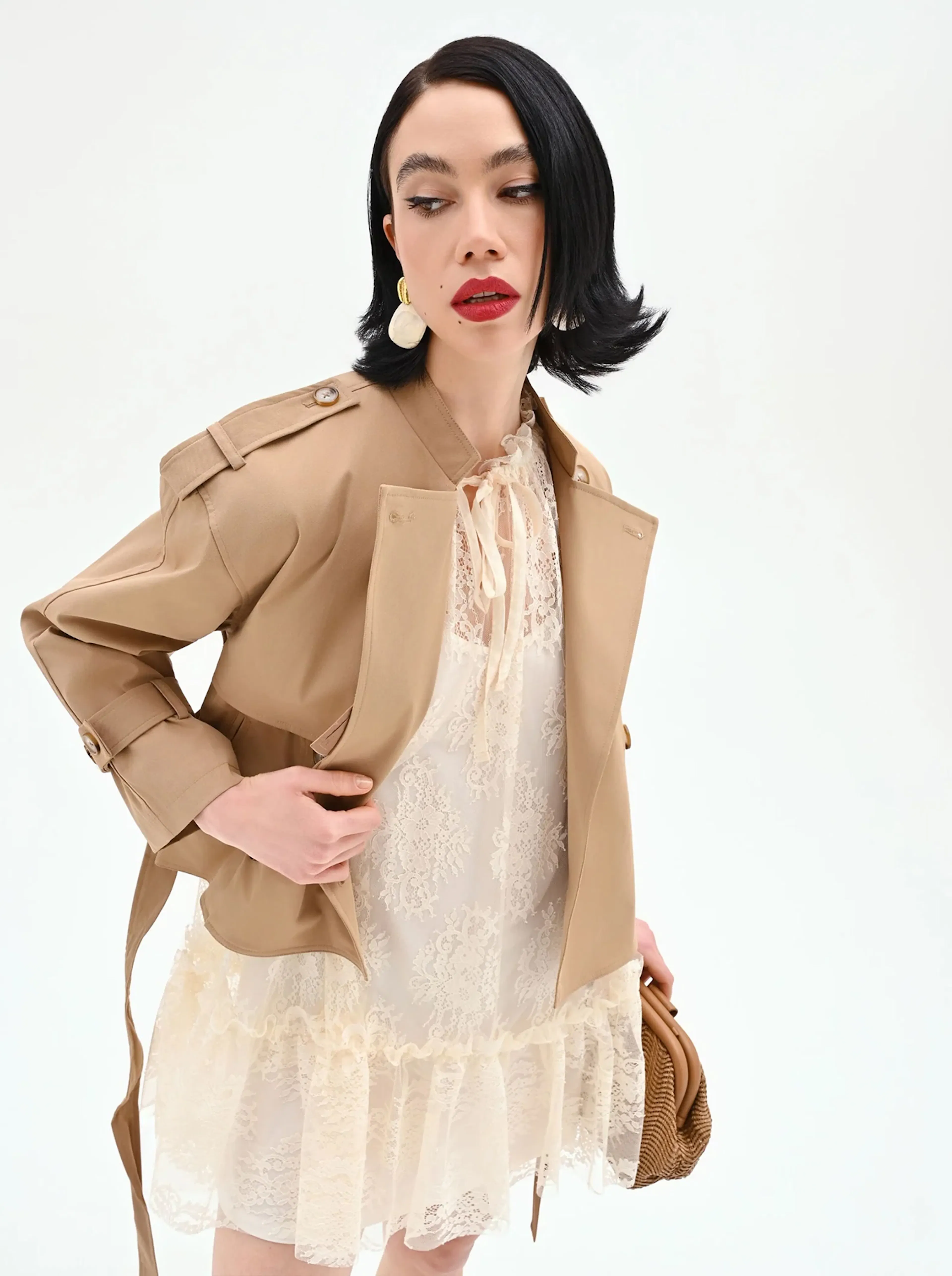 A woman with black hair poses confidently in a beige trench coat over a cream lace dress. She wears large hoop earrings and bold red lipstick.